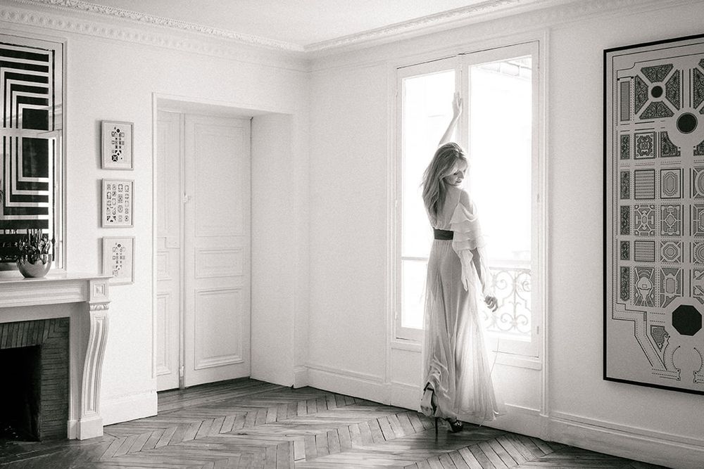 Minimalist interior scene with figure in white dress silhouetted against bright Parisian windows.