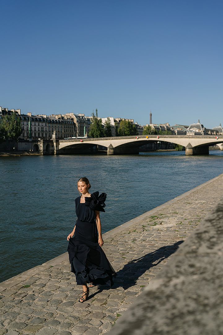 Black dress silhouette walking along Seine River with historic Paris bridge and architecture in background.