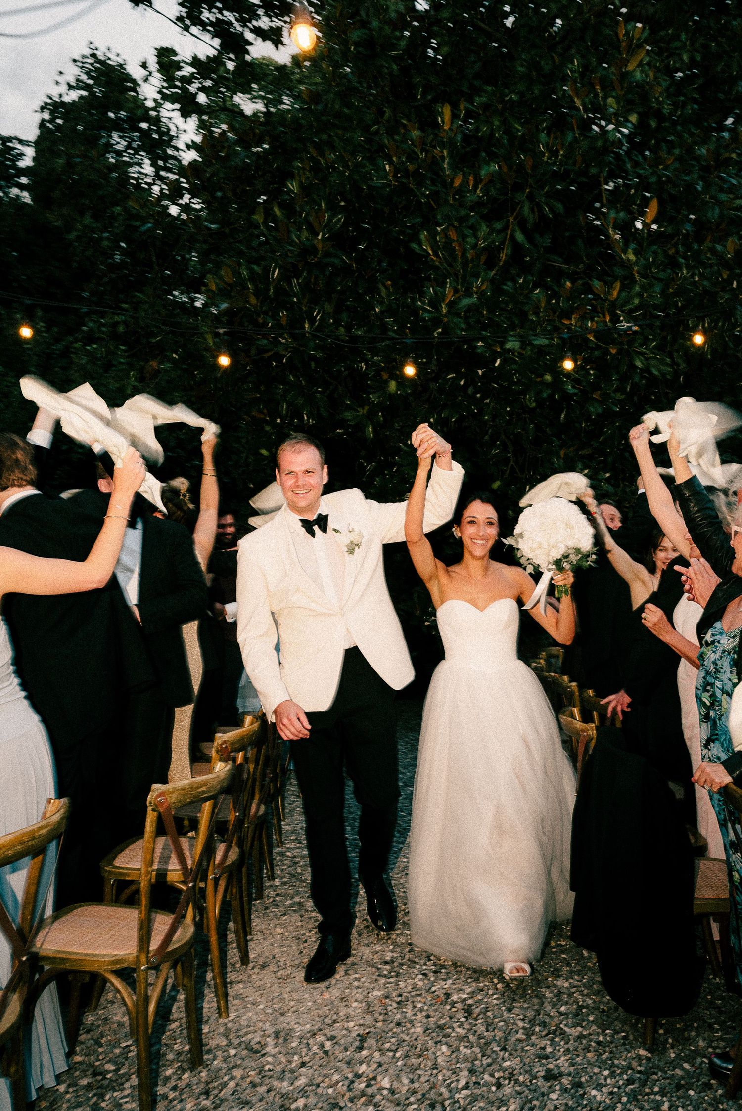 Wedding celebration at night with white doves being released as guests gather around on a gravel path with string lights above.