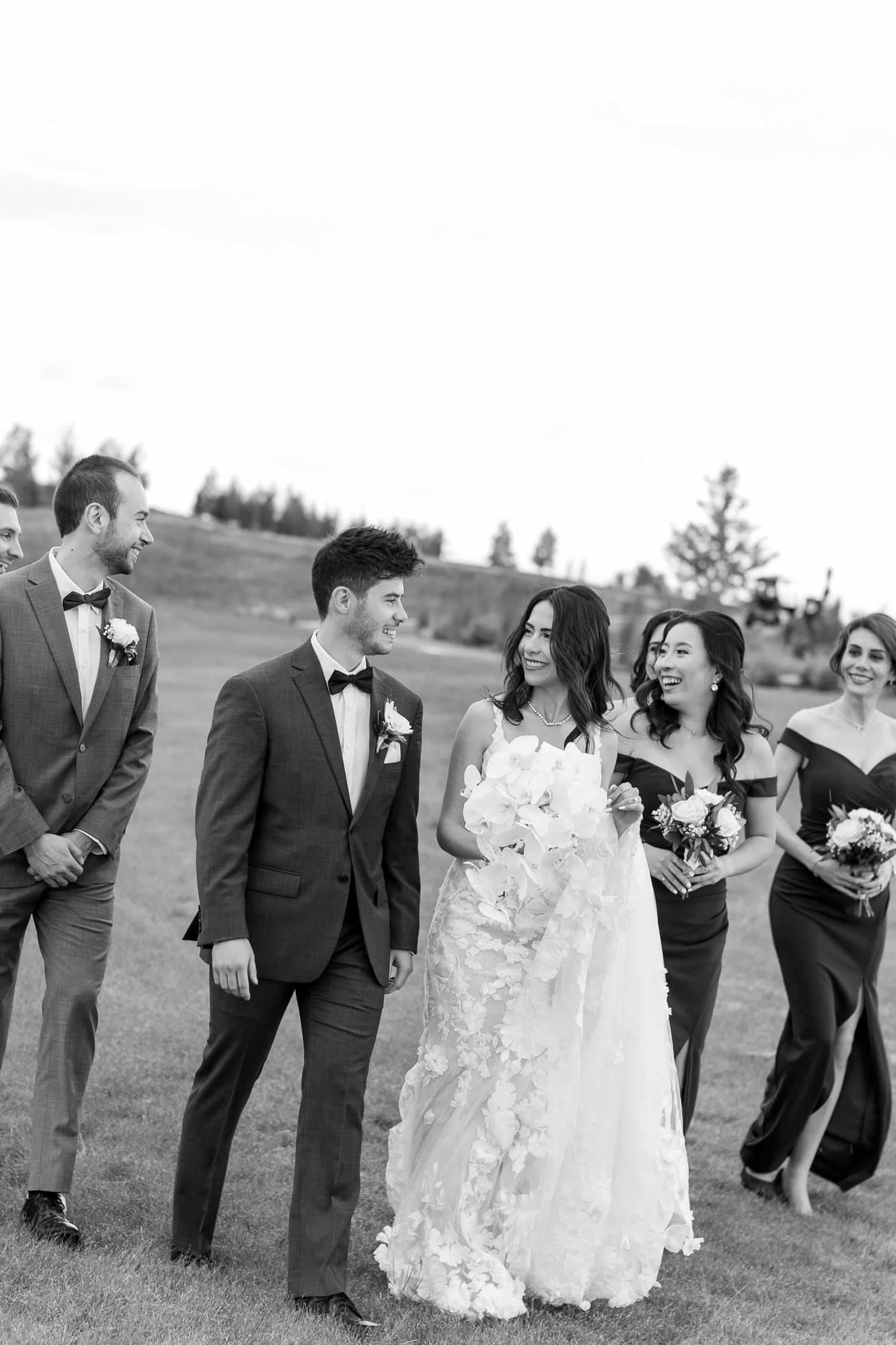 Wedding party laughs together while standing in a grassy field on a sunny day.