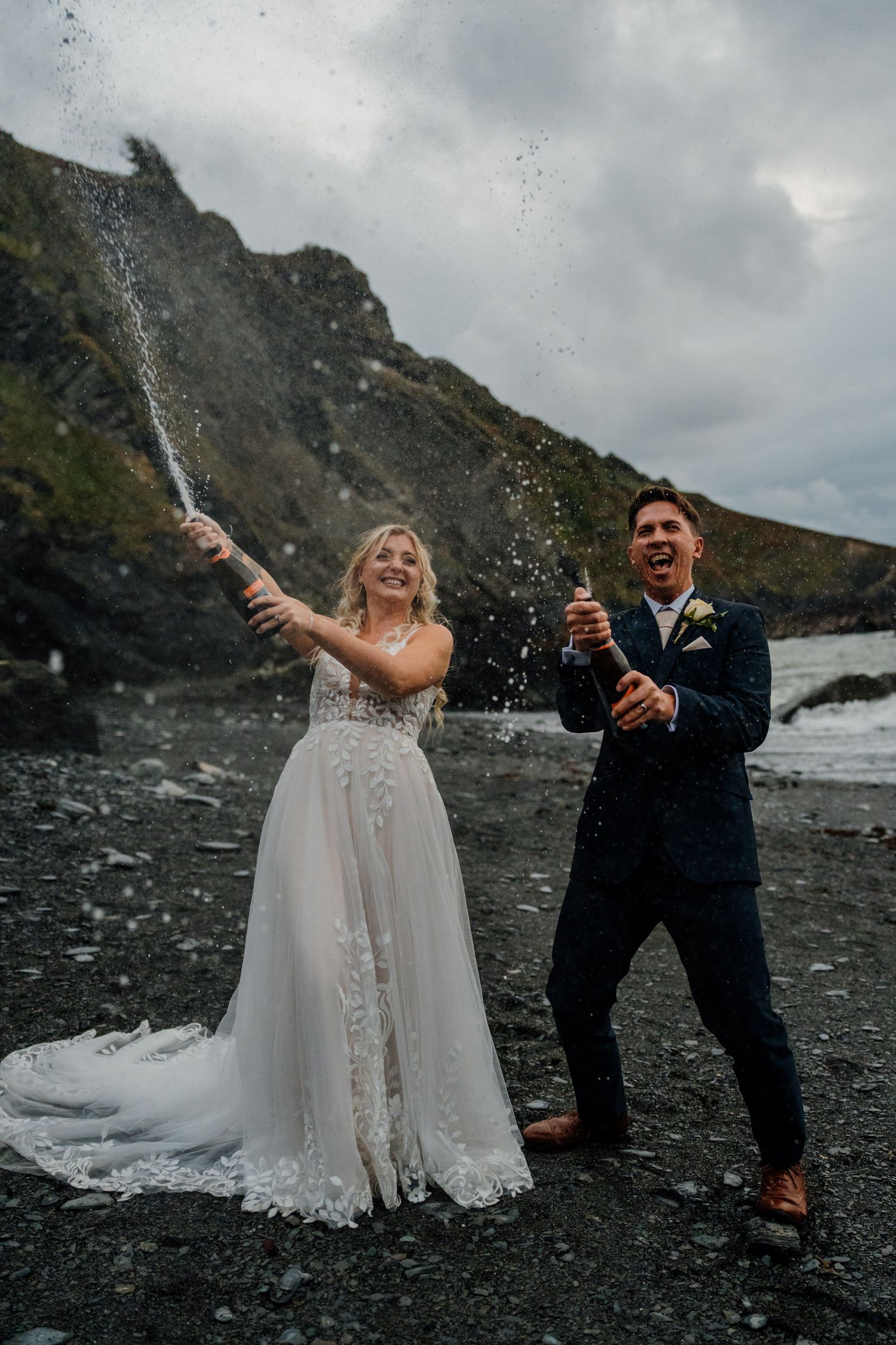A couple celebrates on a dramatic black beach coastline by popping champagne bottles against a moody sky.