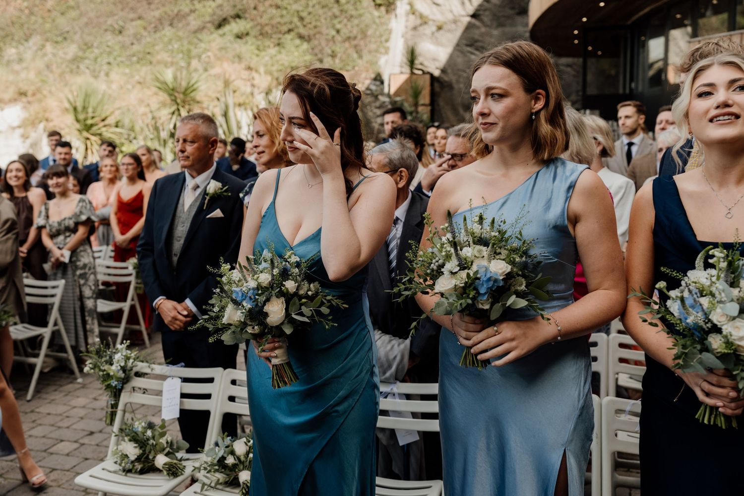 Wedding party in coordinated blue dresses holding floral bouquets outdoors.
