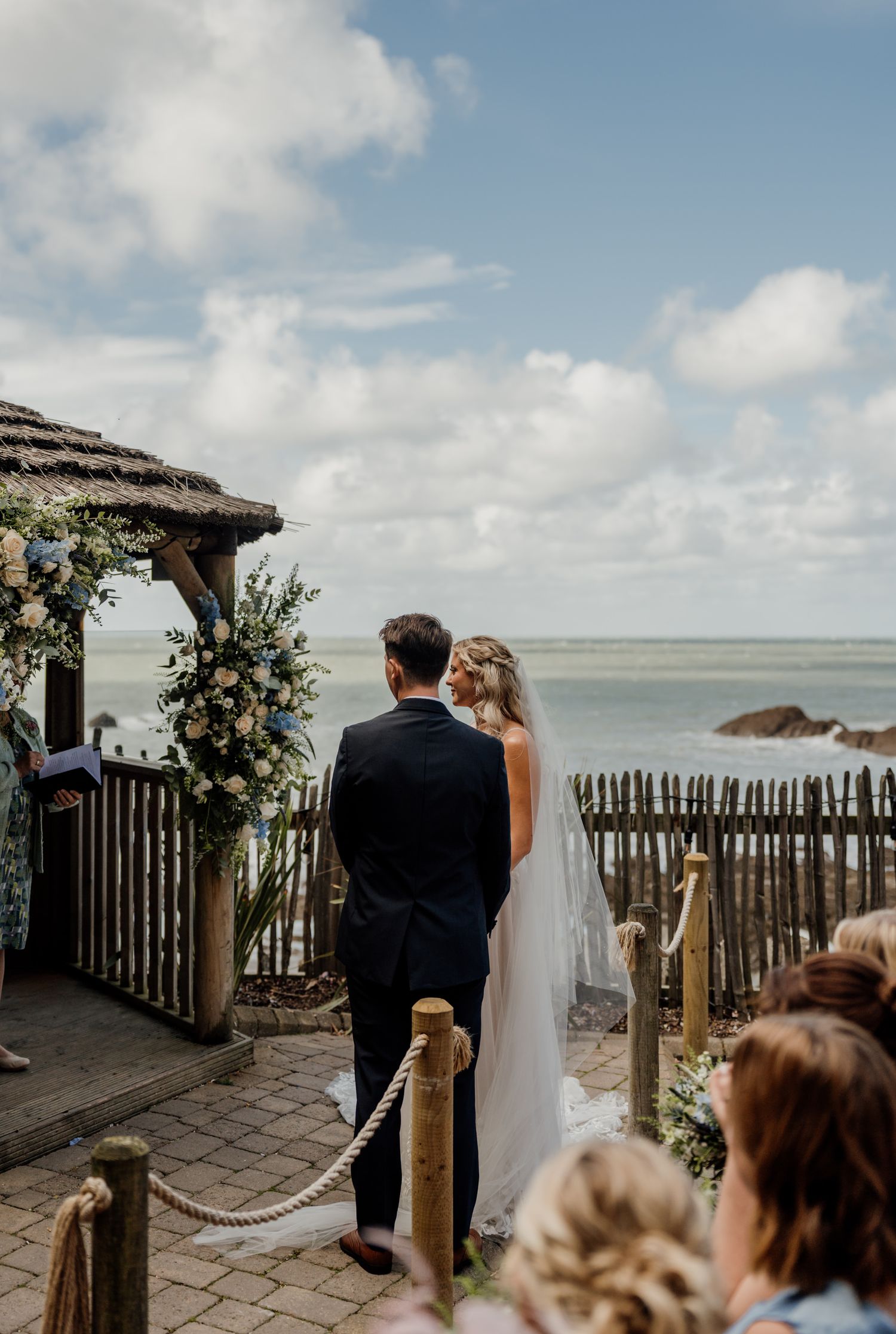 Coastal wedding ceremony with couple standing under flower-adorned gazebo overlooking the ocean.