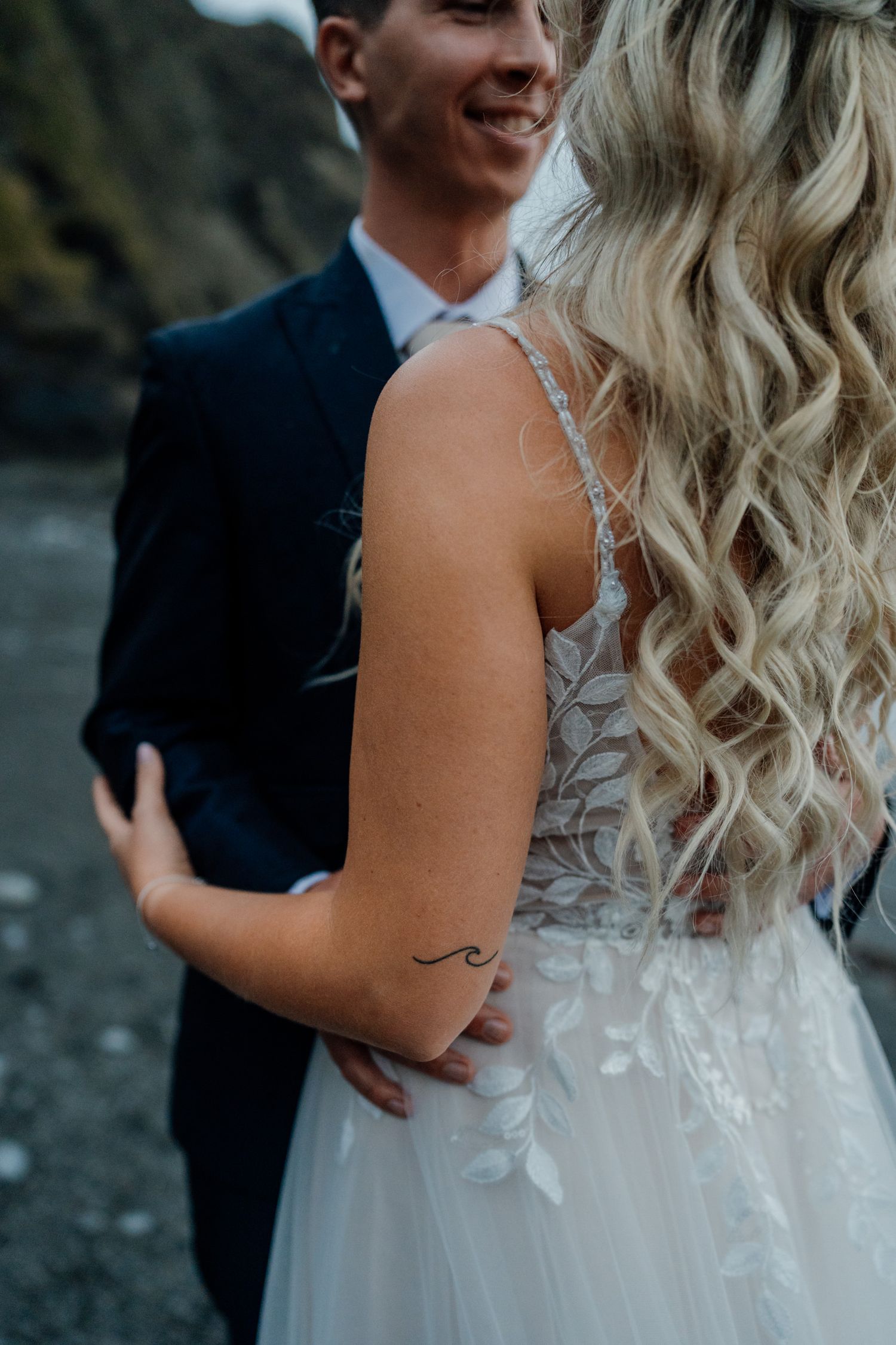 Close-up of bride's blonde curls and lace wedding dress detail with small tattoo visible on arm.