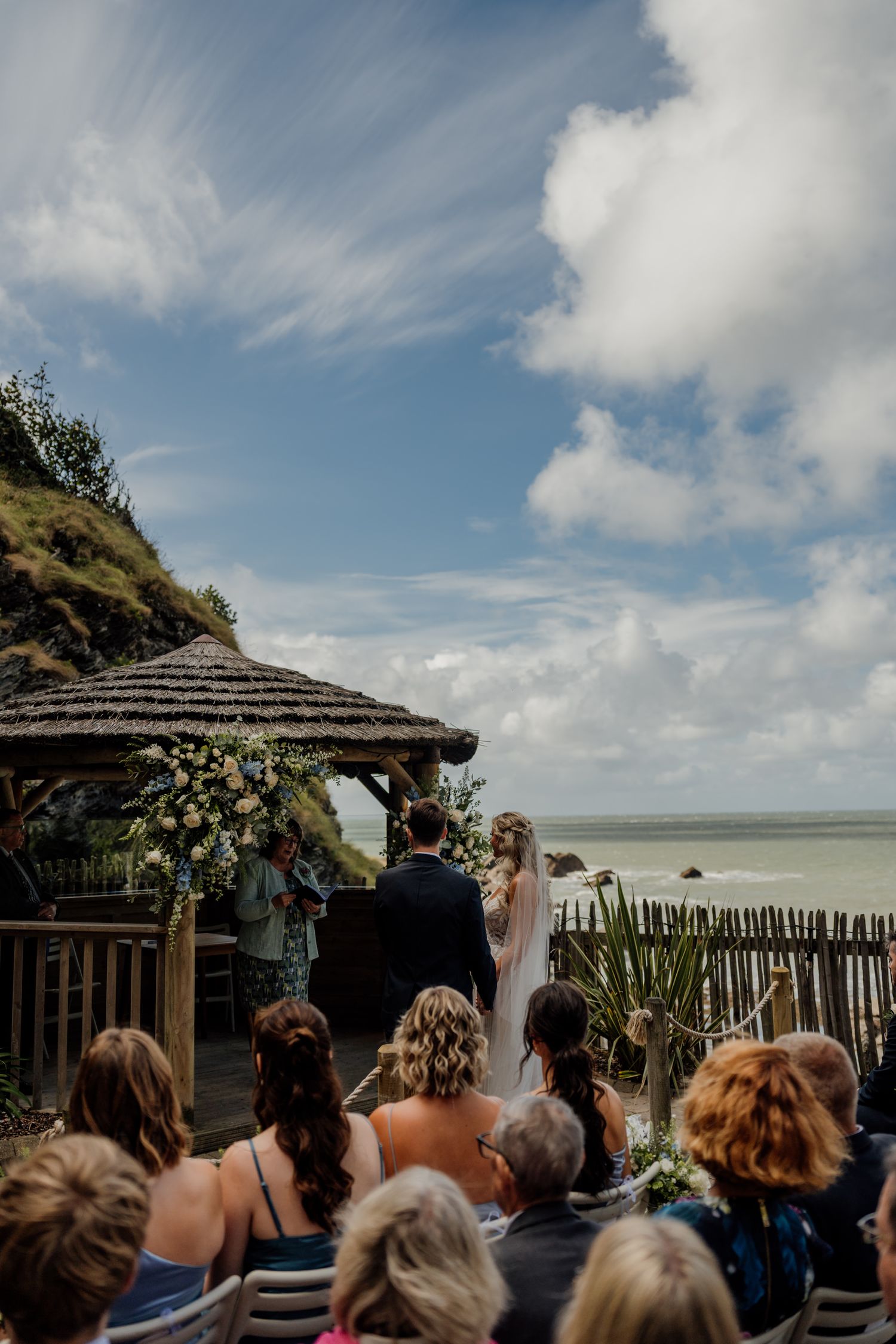 Outdoor coastal wedding ceremony setup with guests seated under blue skies and white clouds at a seaside venue.