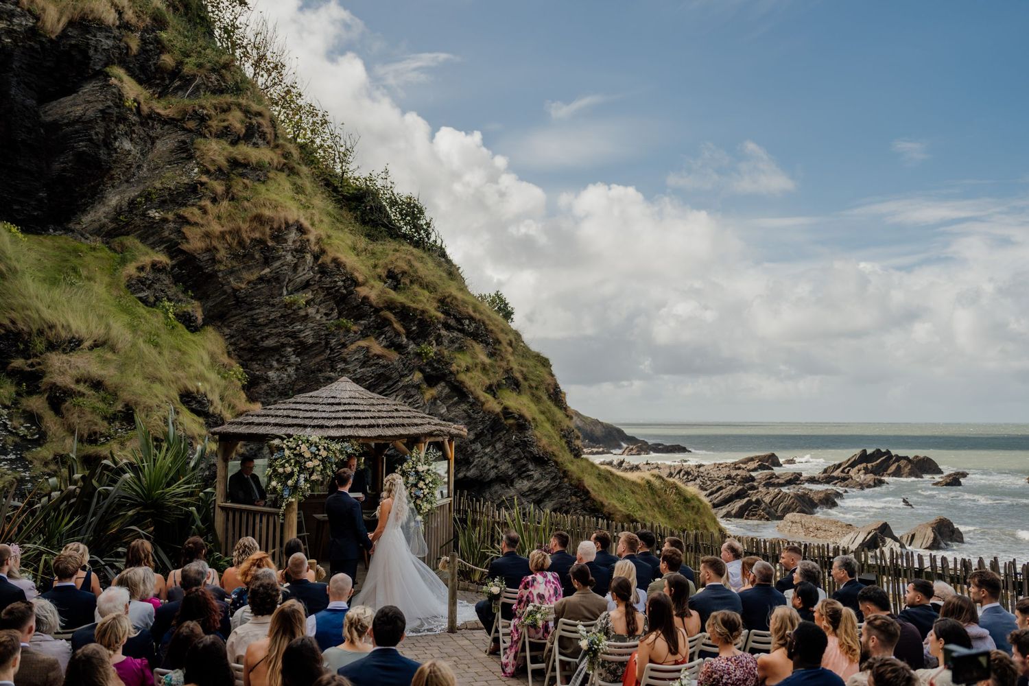 Wedding ceremony taking place at a scenic coastal venue with cliffs and ocean views in the background.