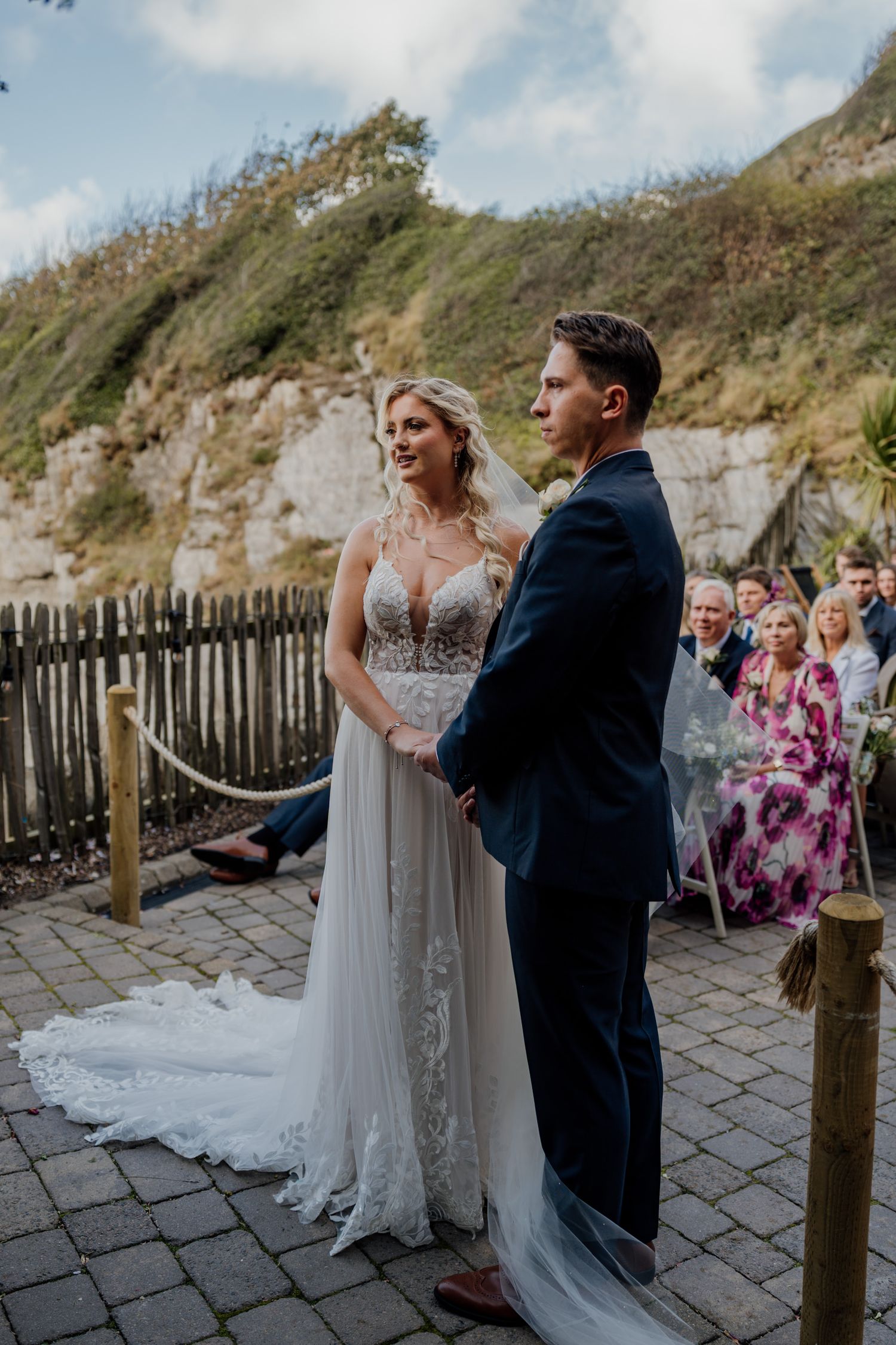 A romantic wedding ceremony unfolds on a coastal boardwalk with guests watching from wooden benches.