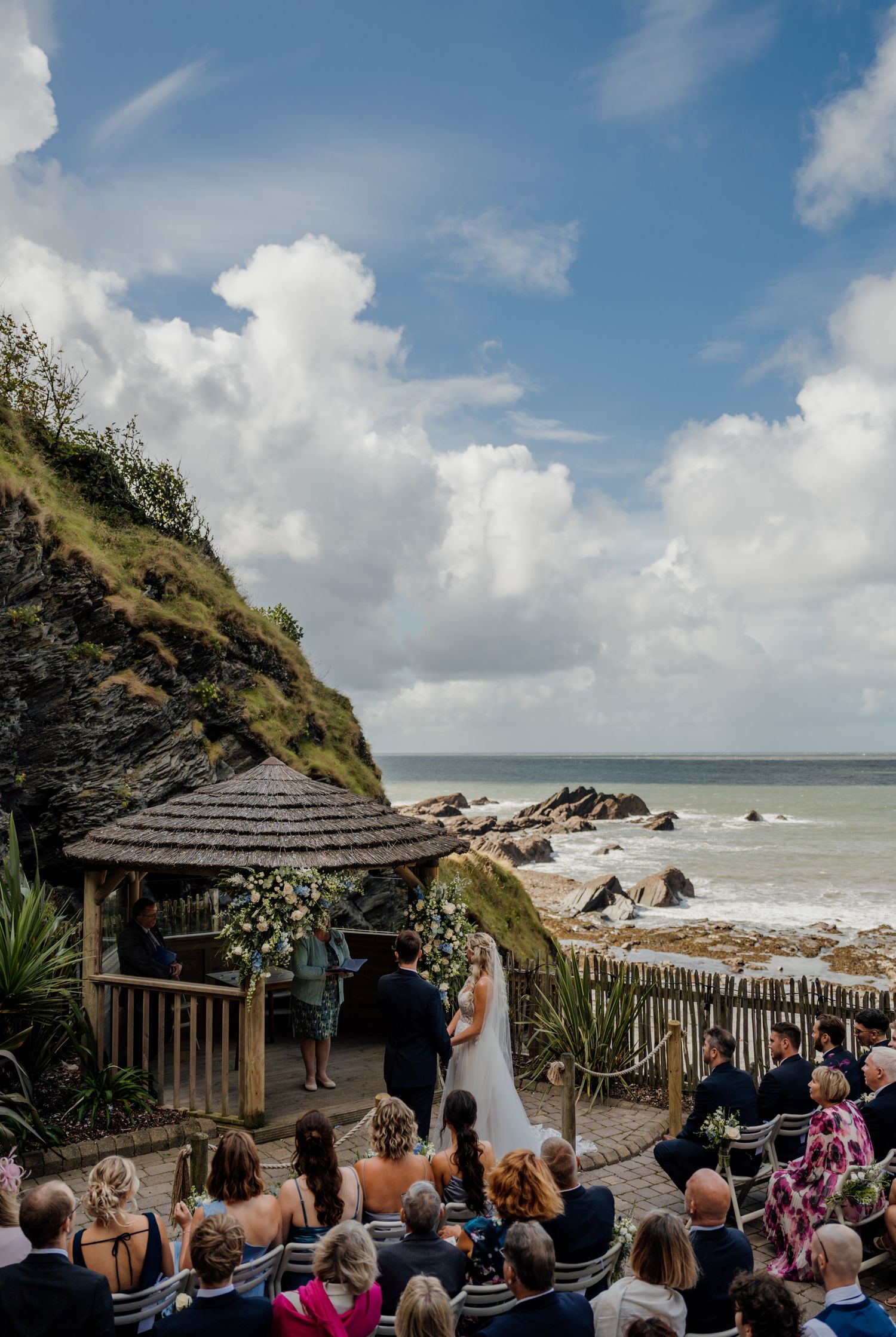 Oceanside wedding ceremony in progress at a clifftop gazebo with guests seated on white chairs.