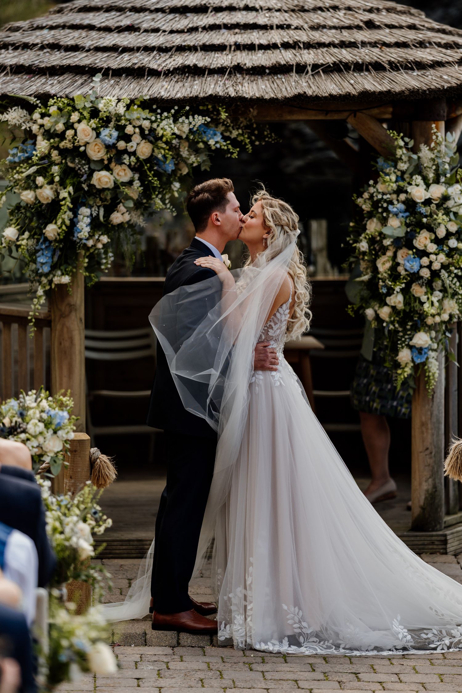 A rustic outdoor wedding venue decorated with white floral arrangements and a thatched roof arbor.