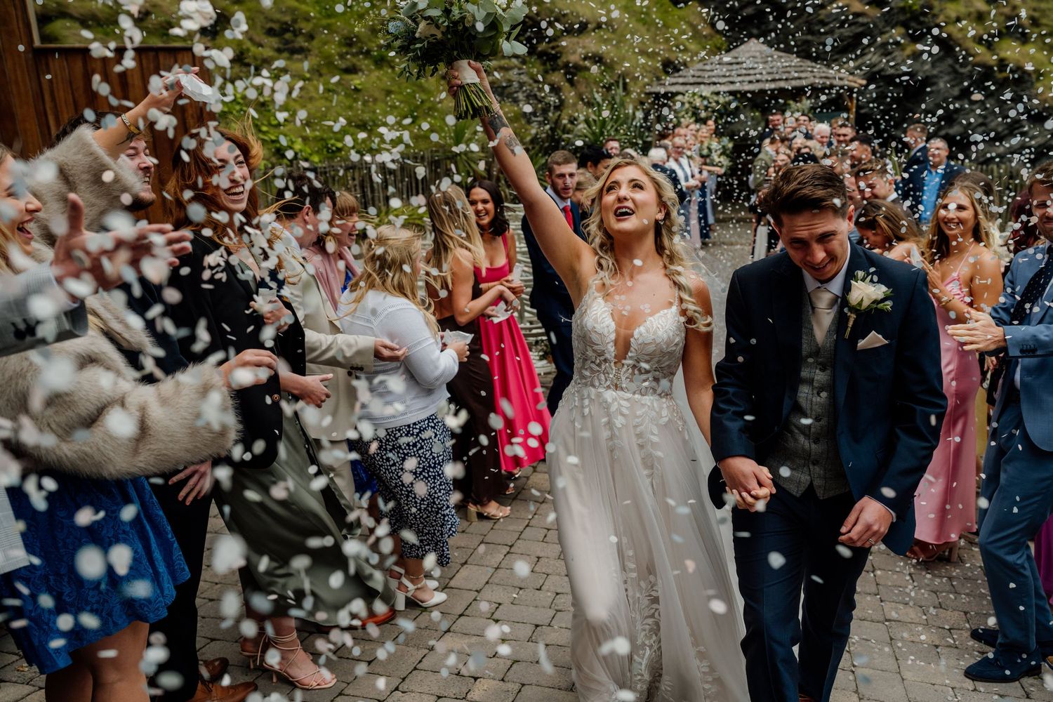 Newlyweds walking through shower of confetti on stone pathway during wedding exit.