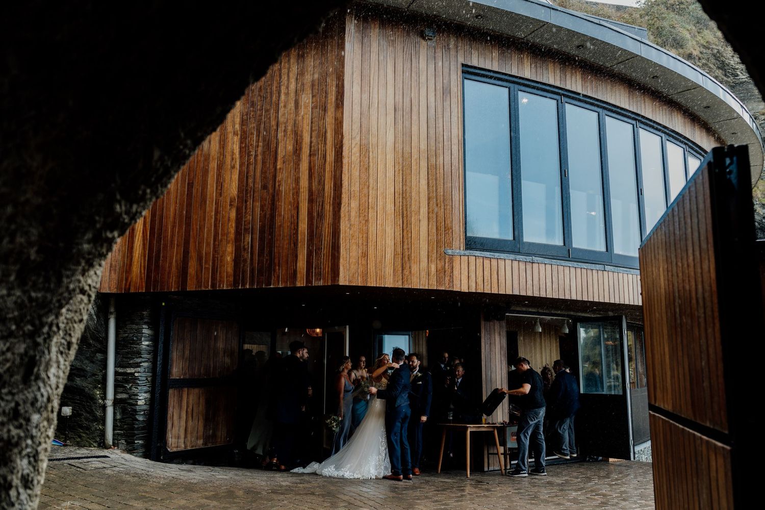 Modern wooden wedding venue with large windows nestled against coastal cliffs.