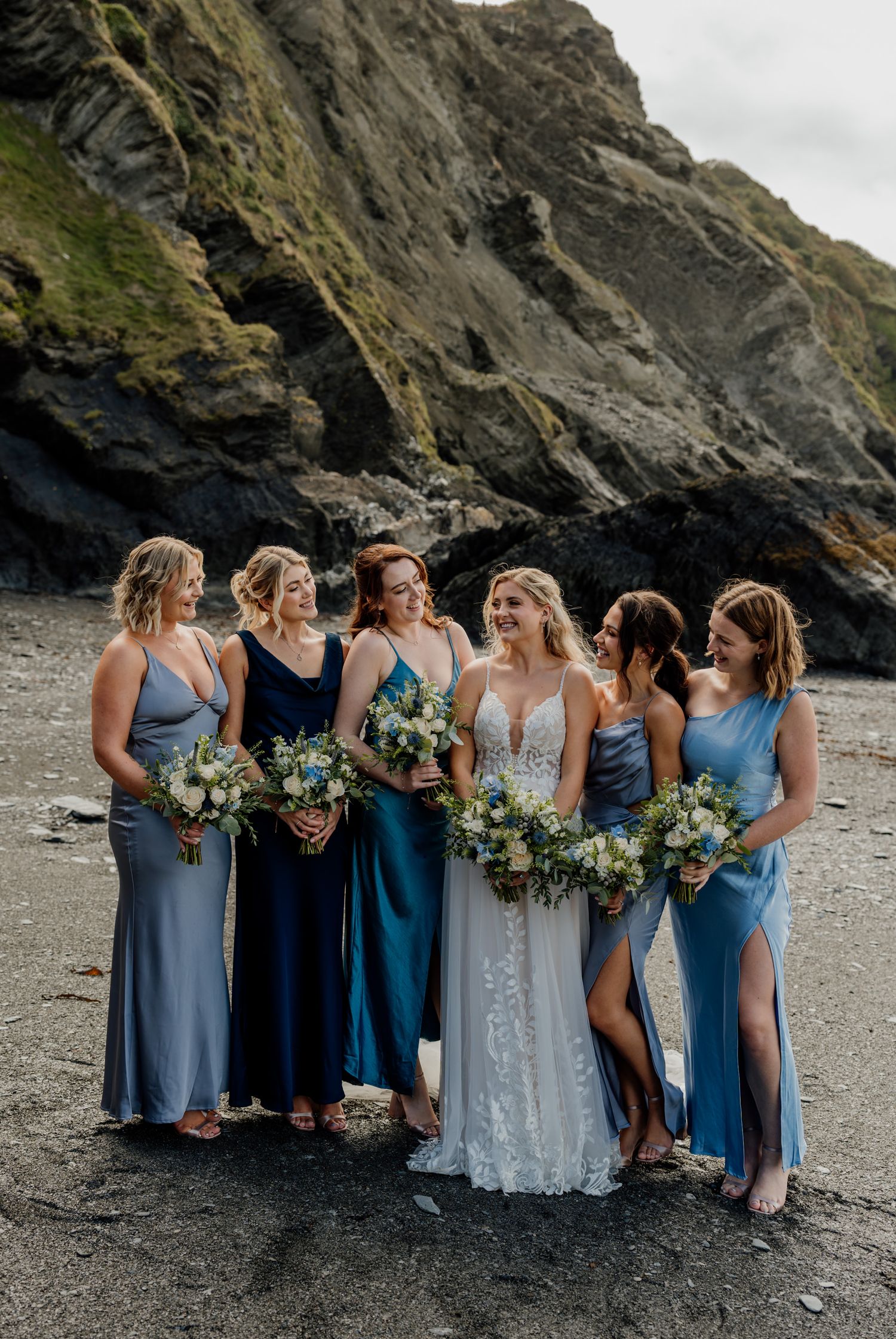 Bridesmaids in varying shades of blue dresses posed together on a rocky beach.
