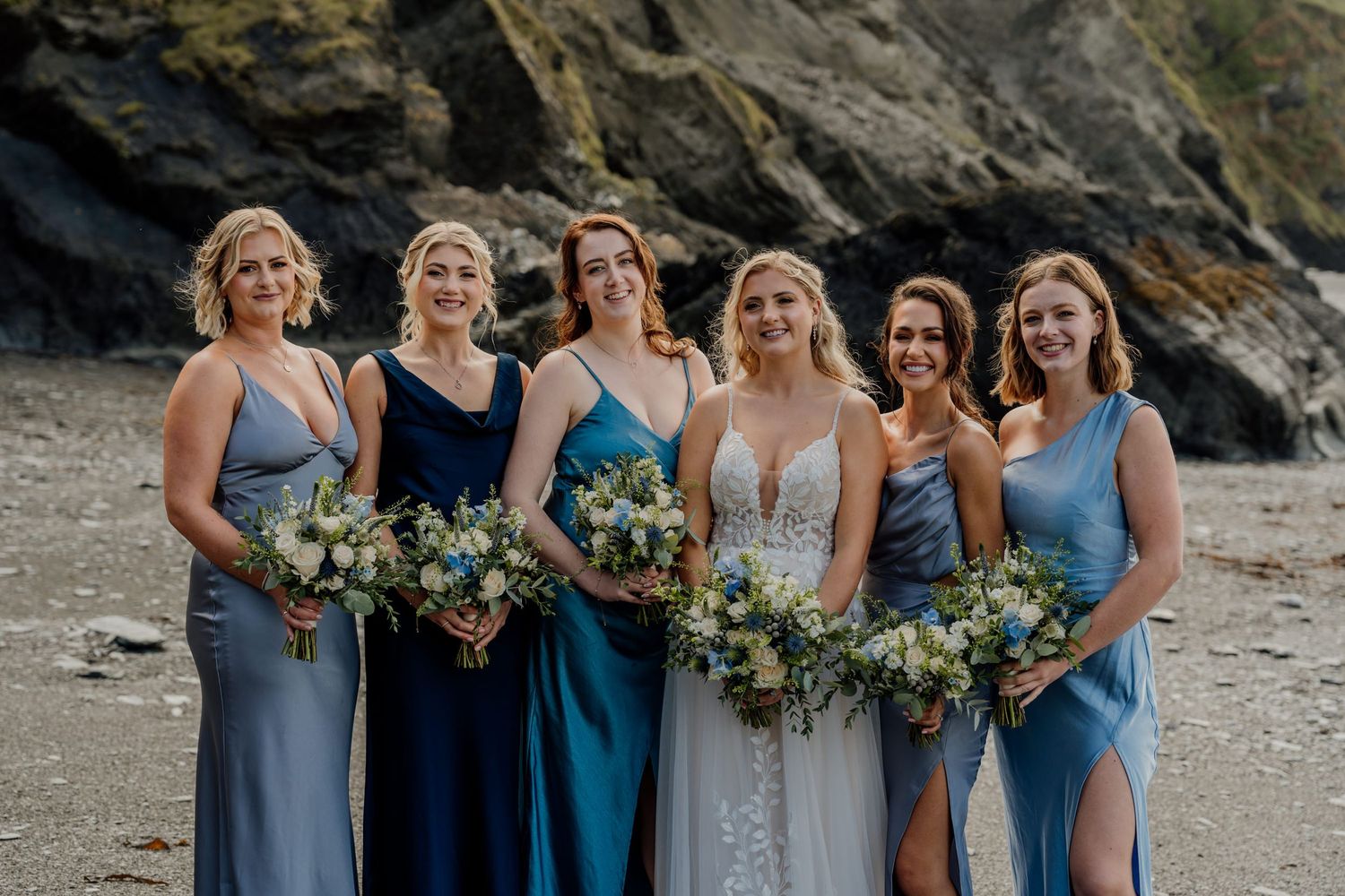 Bridesmaids in blue dresses holding bouquets on a coastal beach.