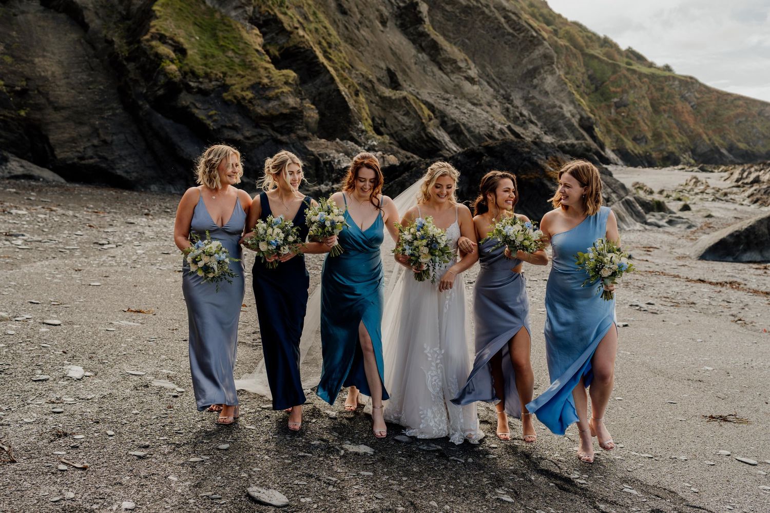 Bridal party in blue dresses posing together on rocky beach.
