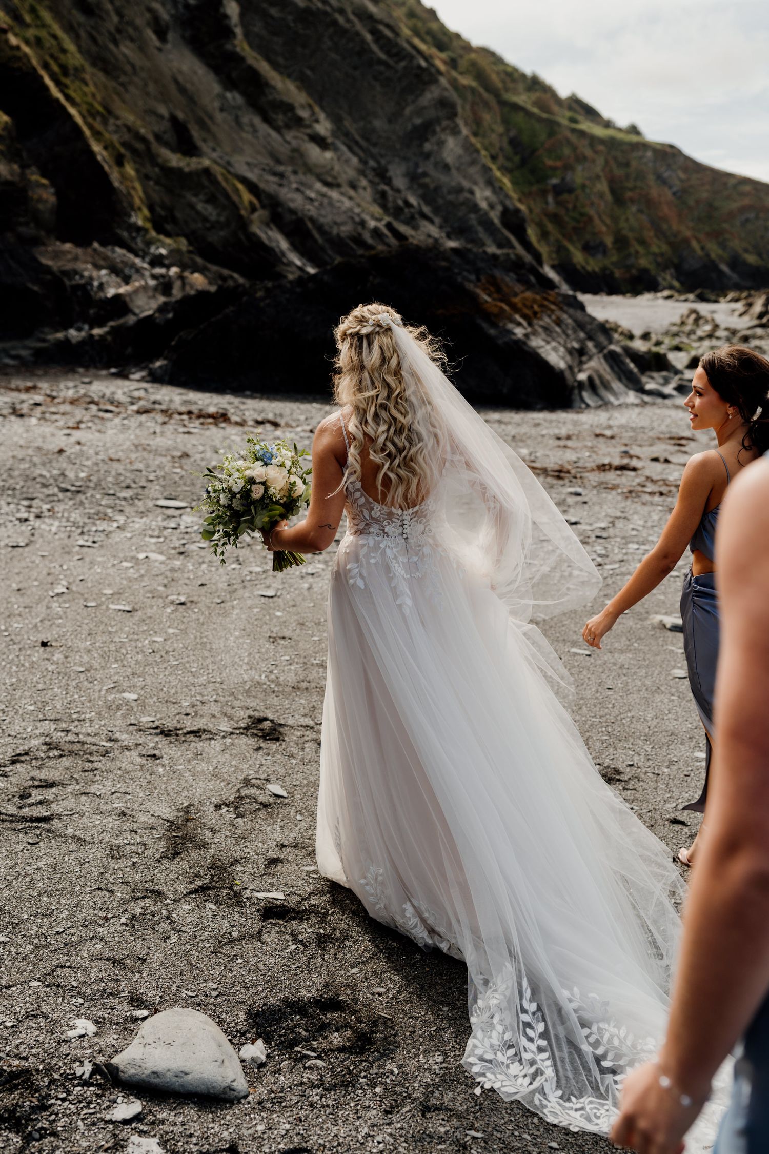 Bride walking along a dramatic rocky beach with flowing veil and bouquet.