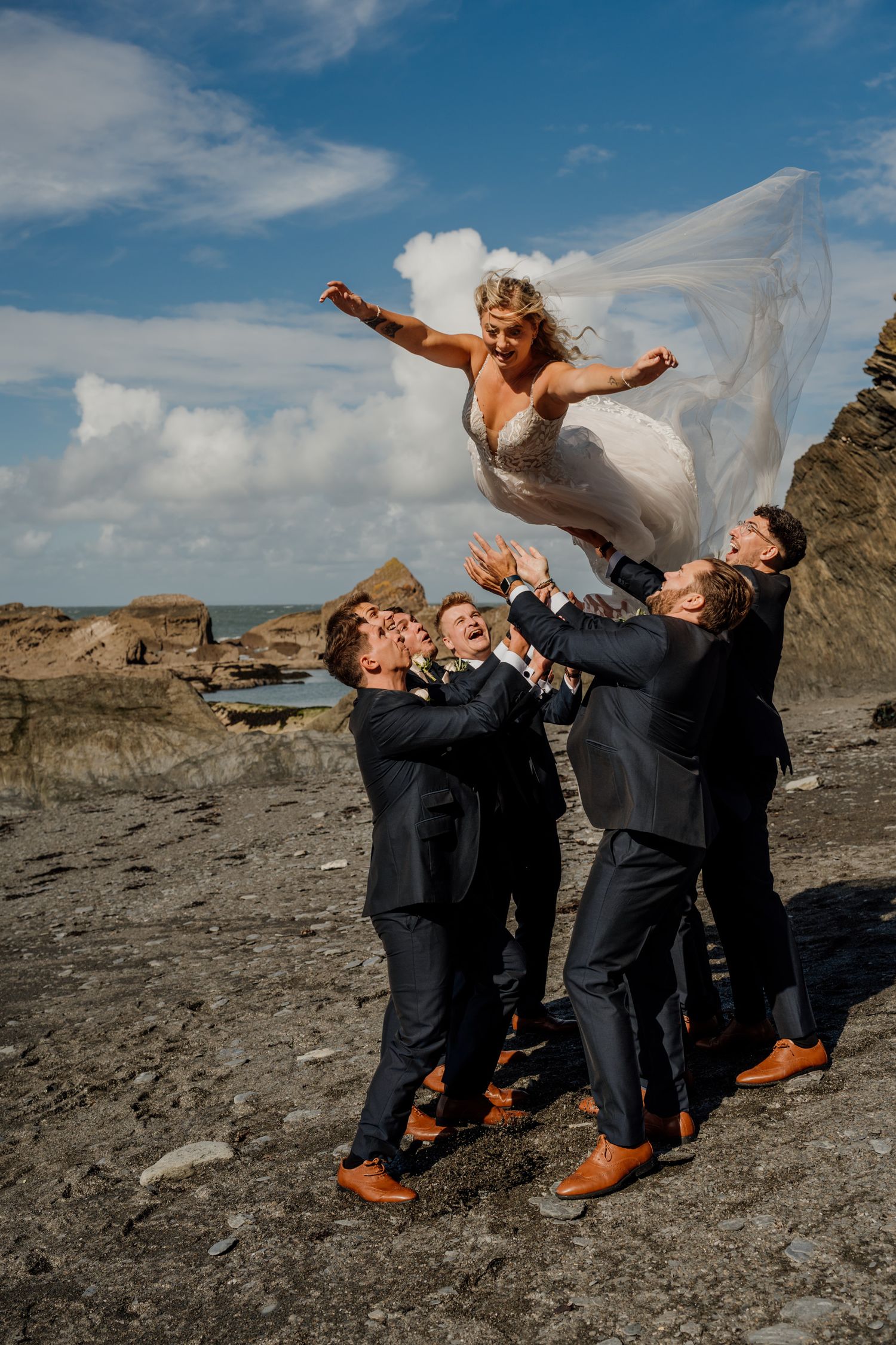 Fun wedding party photo with groomsmen tossing the bride on rocky coastal backdrop.