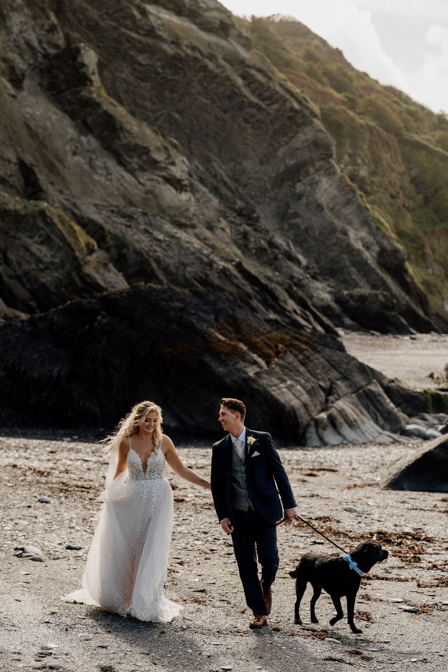 Wedding couple walking with their black dog on a remote beach with rocky cliffs.