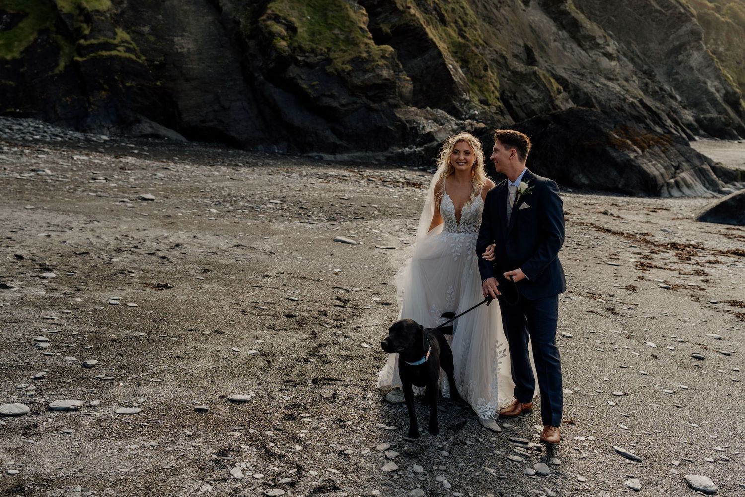 Couple with their dog posing on rocky beach in wedding attire.