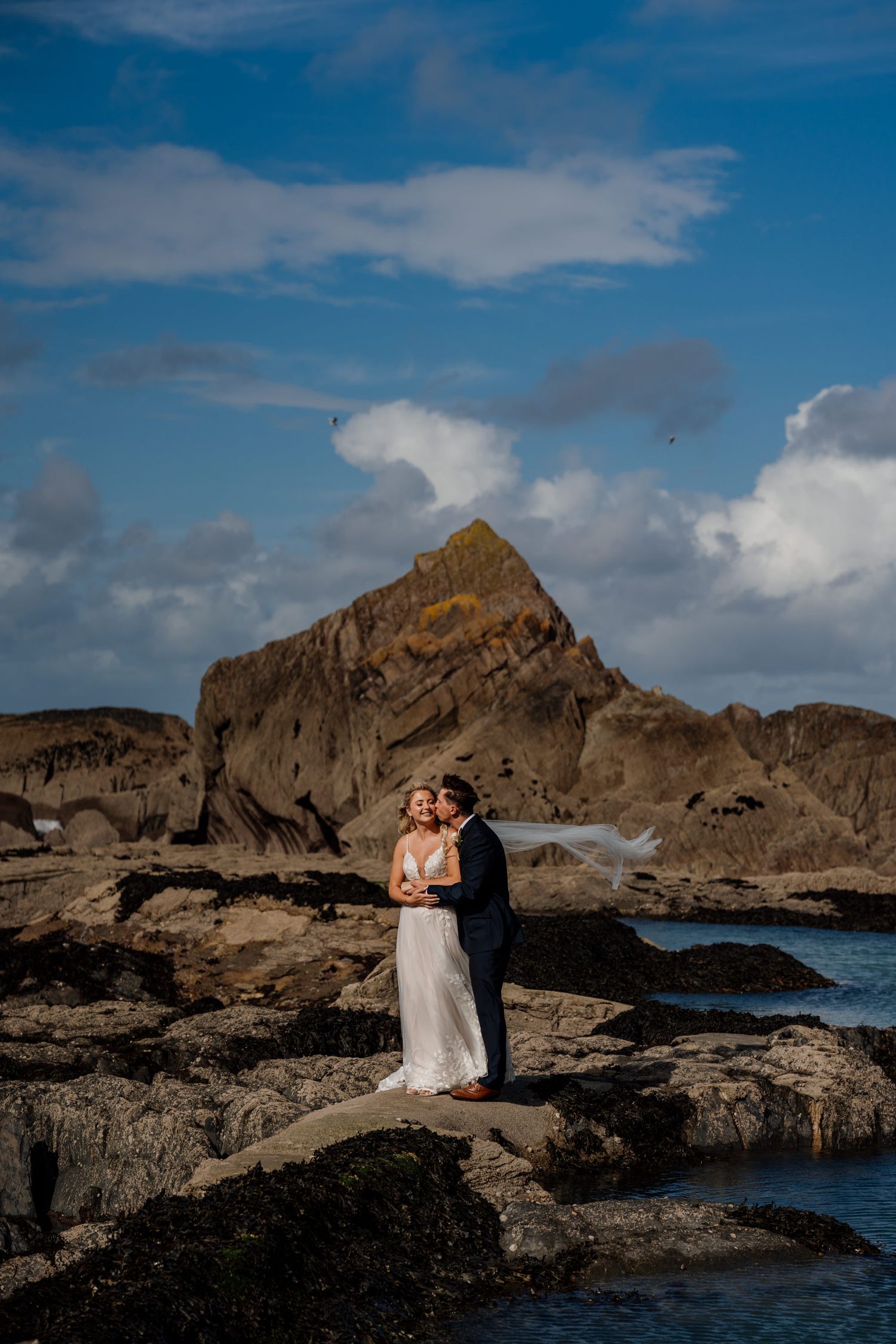Dramatic coastal wedding photos captured against rugged rocky cliffs and blue ocean waters.