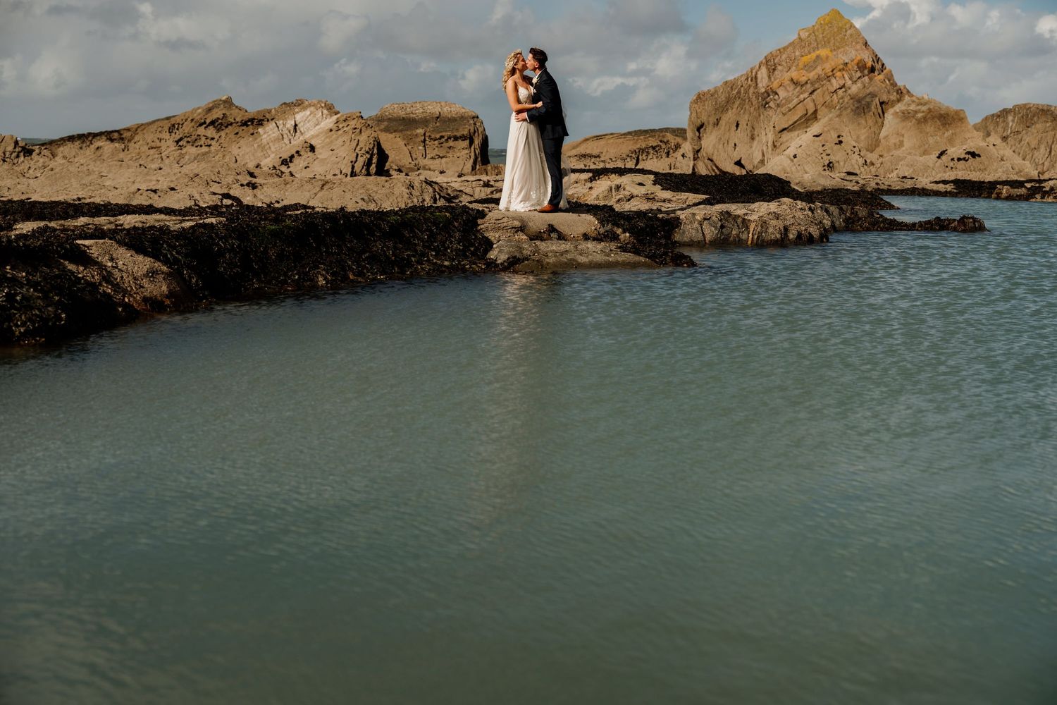 Wedding photography session on rocky coastal outcrop with turquoise water and sandy cliffs.