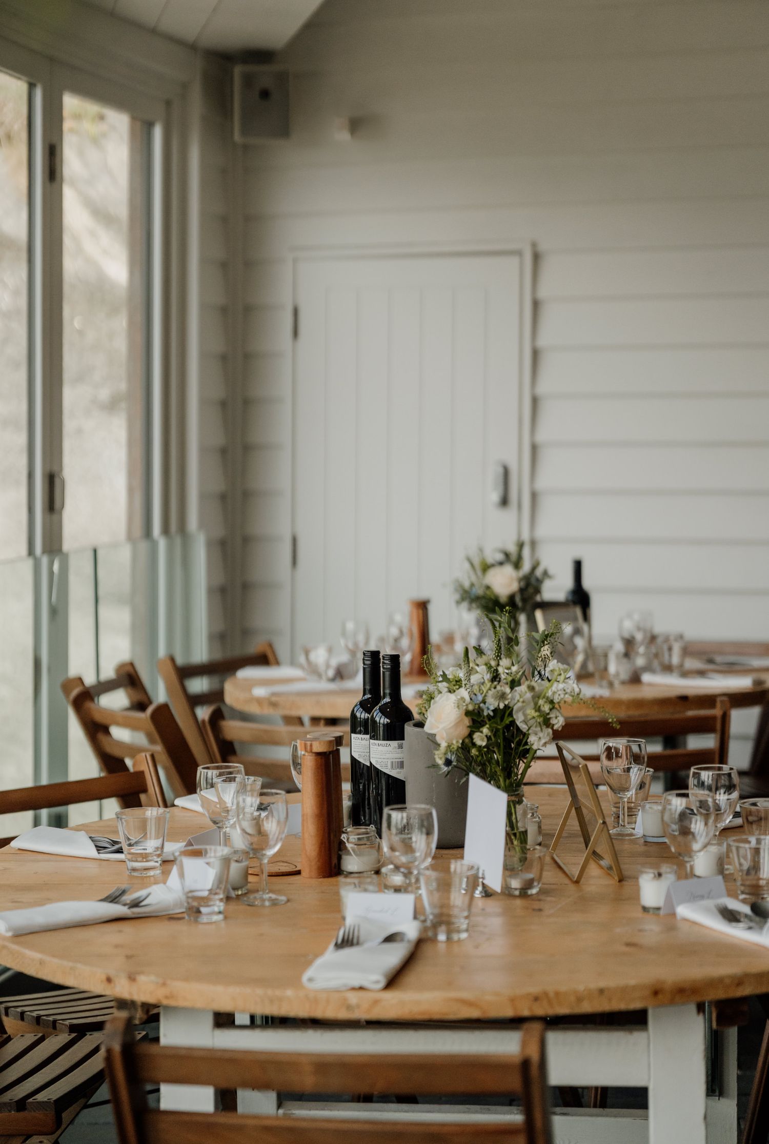 Rustic wedding reception tables set with wooden chairs and simple elegant decor in a bright white room.