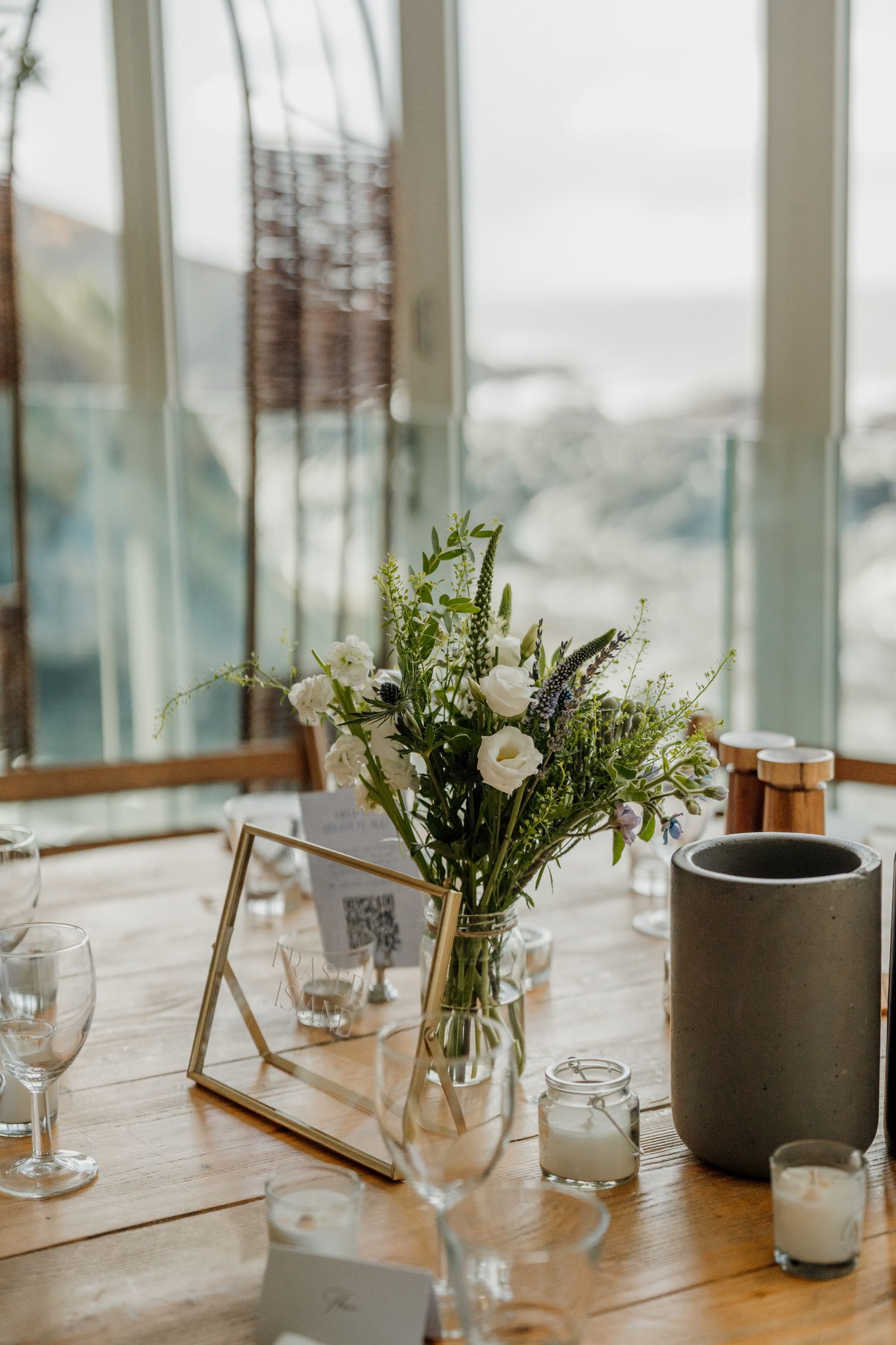 Elegant table setting with fresh white floral centerpiece in vase, wine glasses and candles near large windows with natural light.