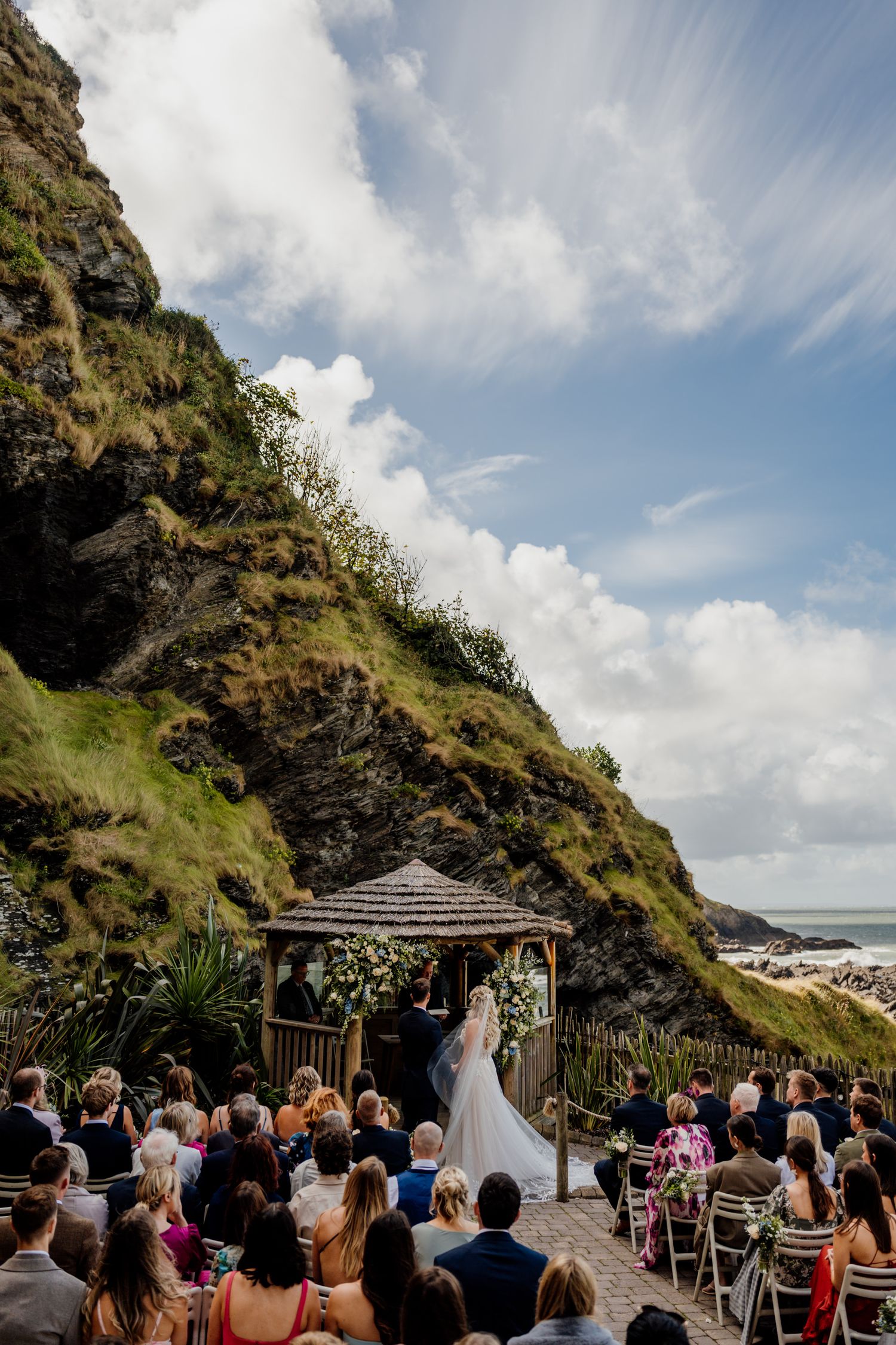 Coastal wedding ceremony taking place in a gazebo overlooking the ocean with guests seated on white chairs.