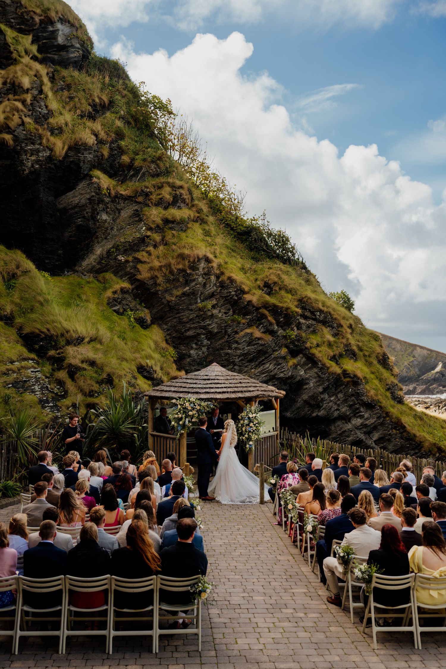 An outdoor wedding ceremony setup on a stone patio with dramatic coastal cliffs and blue sky backdrop.