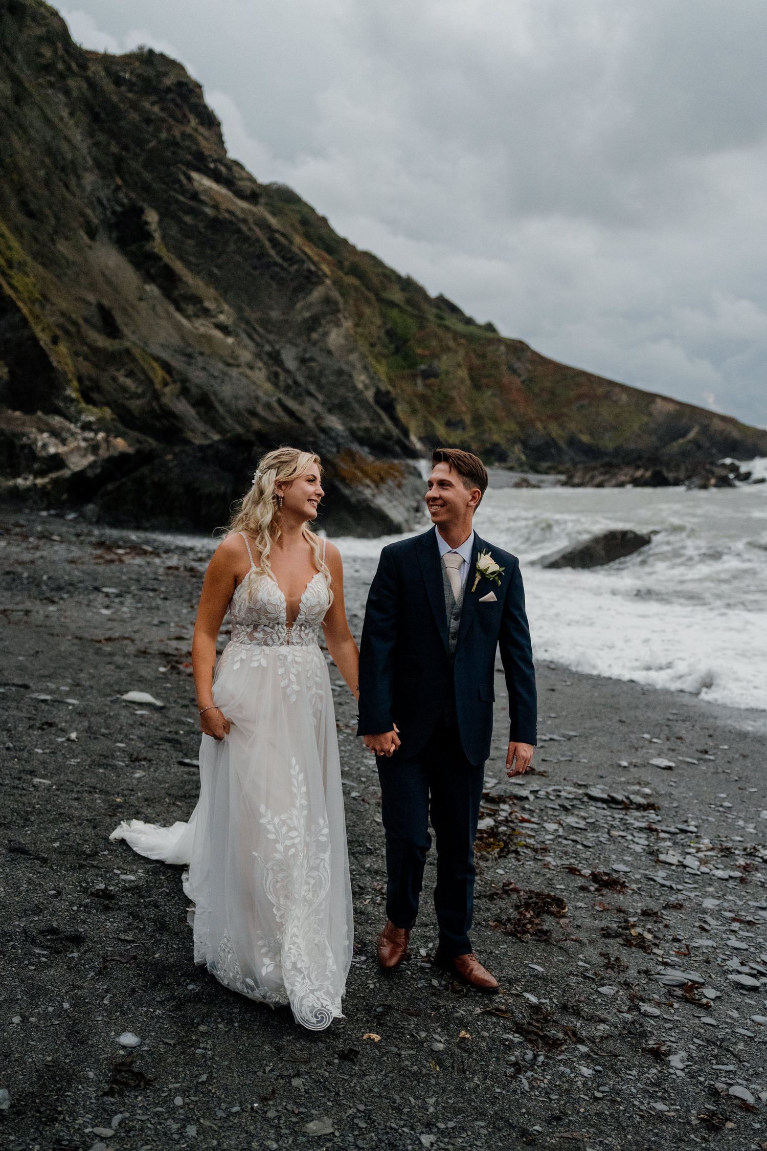 Newlyweds walk hand in hand along a moody black sand beach with rocky cliffs towering behind them.
