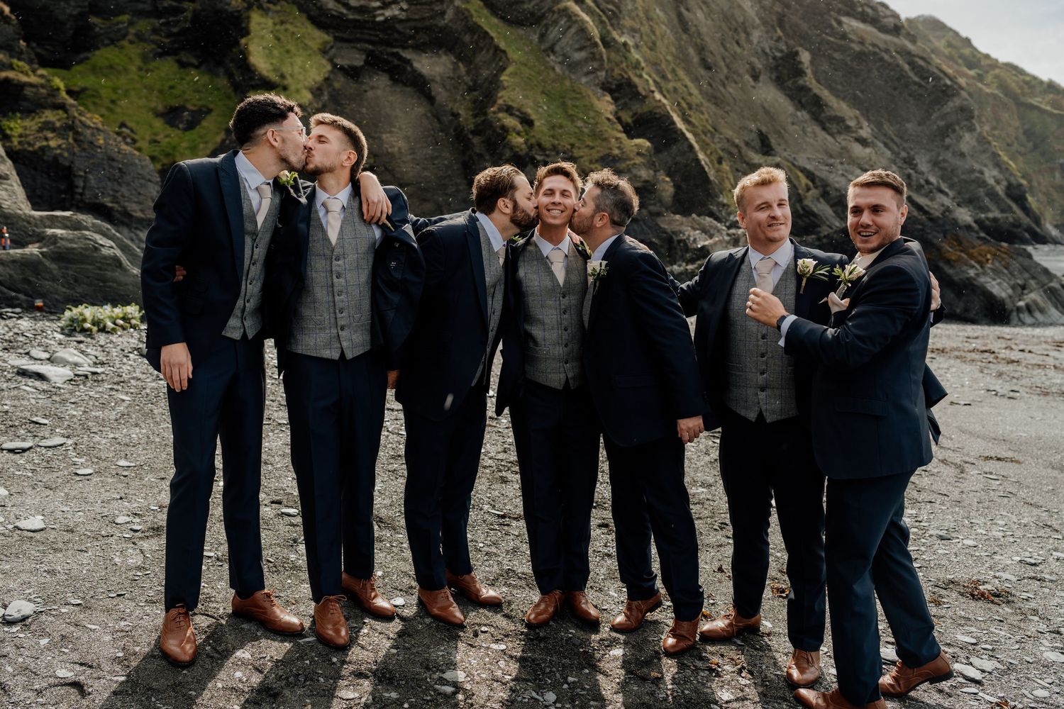 A group wearing dark formal attire poses playfully on rocks along a scenic coastal shoreline.