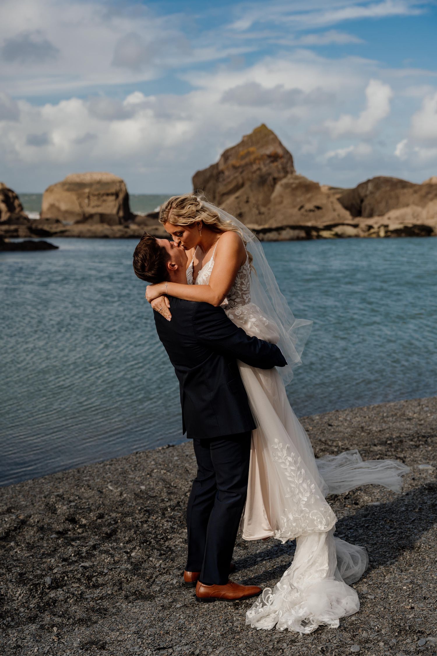 A wedding couple embraces on a rocky beach landscape with dramatic coastal cliffs in the background.