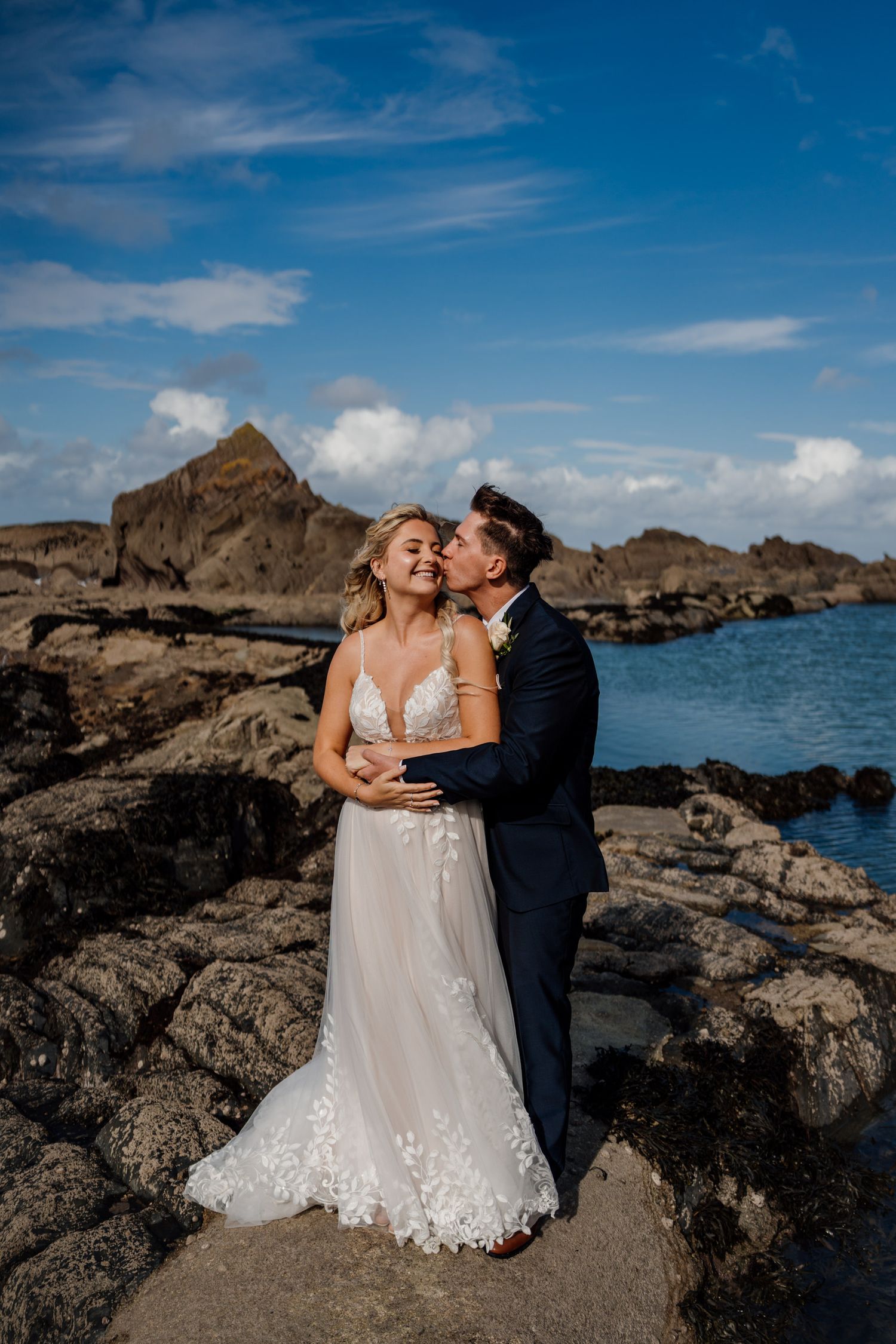 A romantic wedding photoshoot on a rugged beach coastline under a bright blue sky.