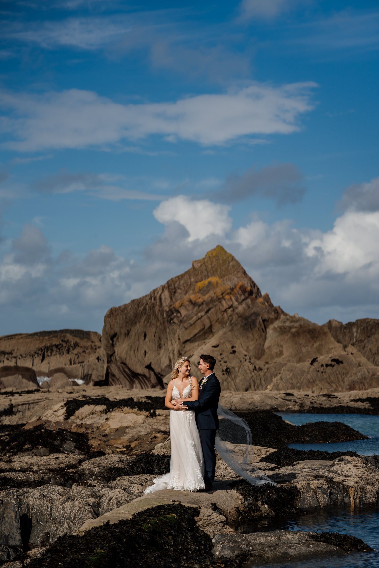 Two figures in wedding attire stand against a scenic coastal backdrop with rocky cliffs and blue skies.