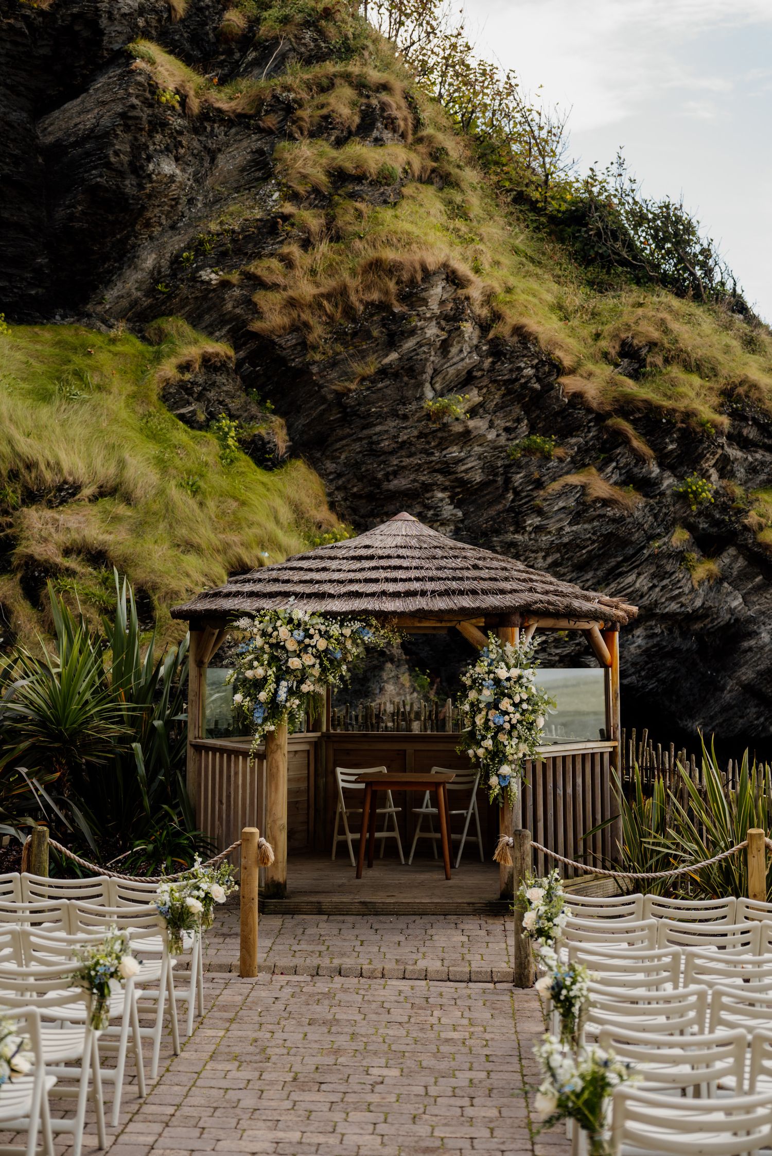 Outdoor wedding ceremony setup with white chairs and floral arrangements under a wooden gazebo against coastal cliffs.