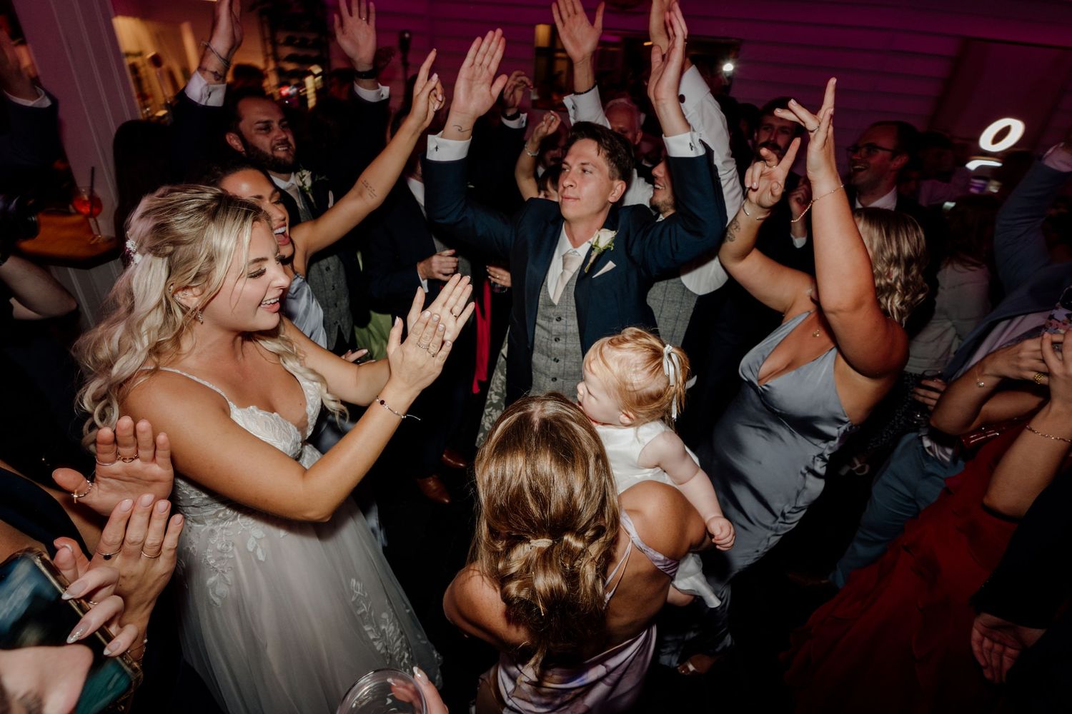 Wedding reception guests dancing energetically with hands raised on a dimly lit dance floor.