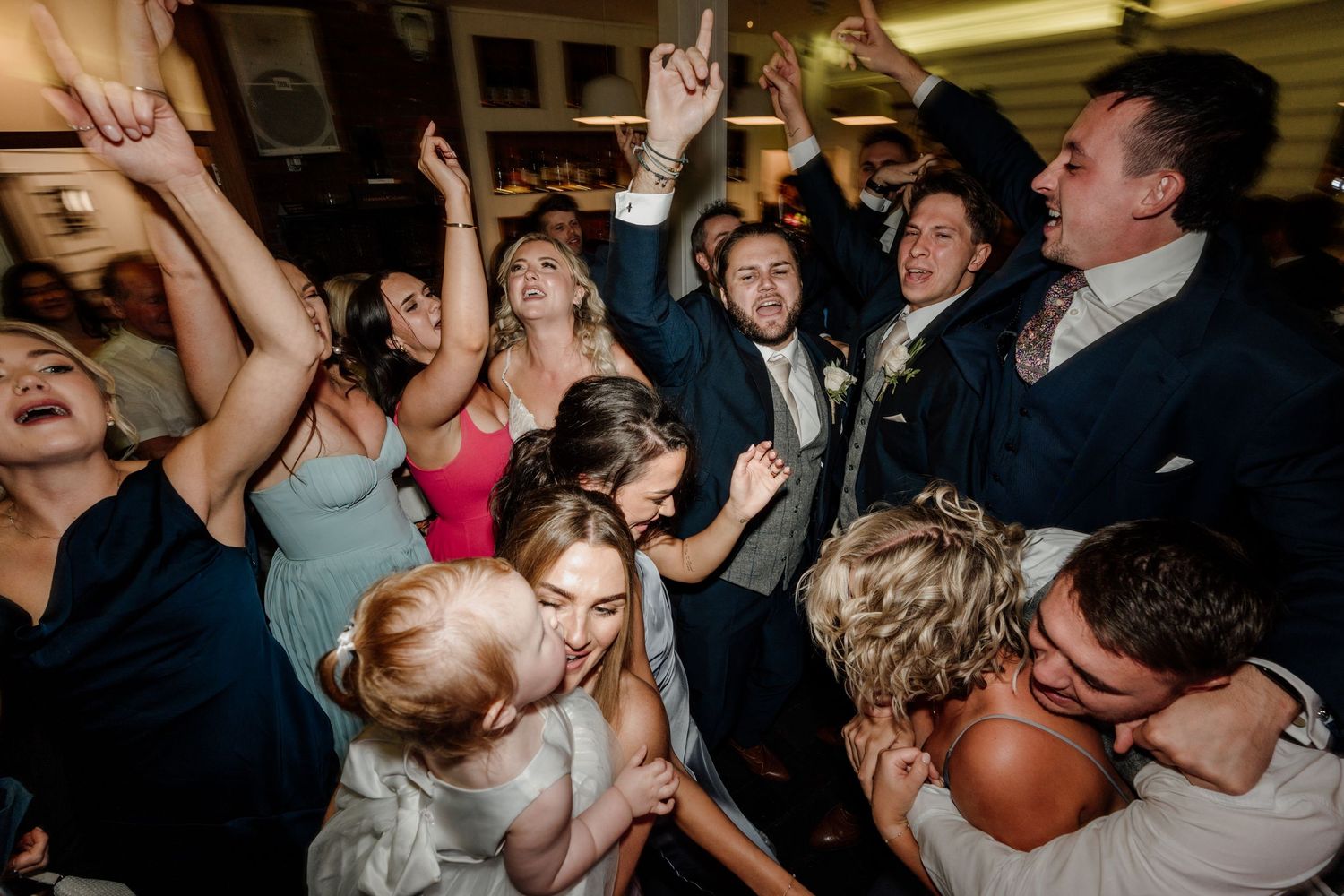 Wedding reception dance party with guests dancing under dimly lit atmospheric lighting.