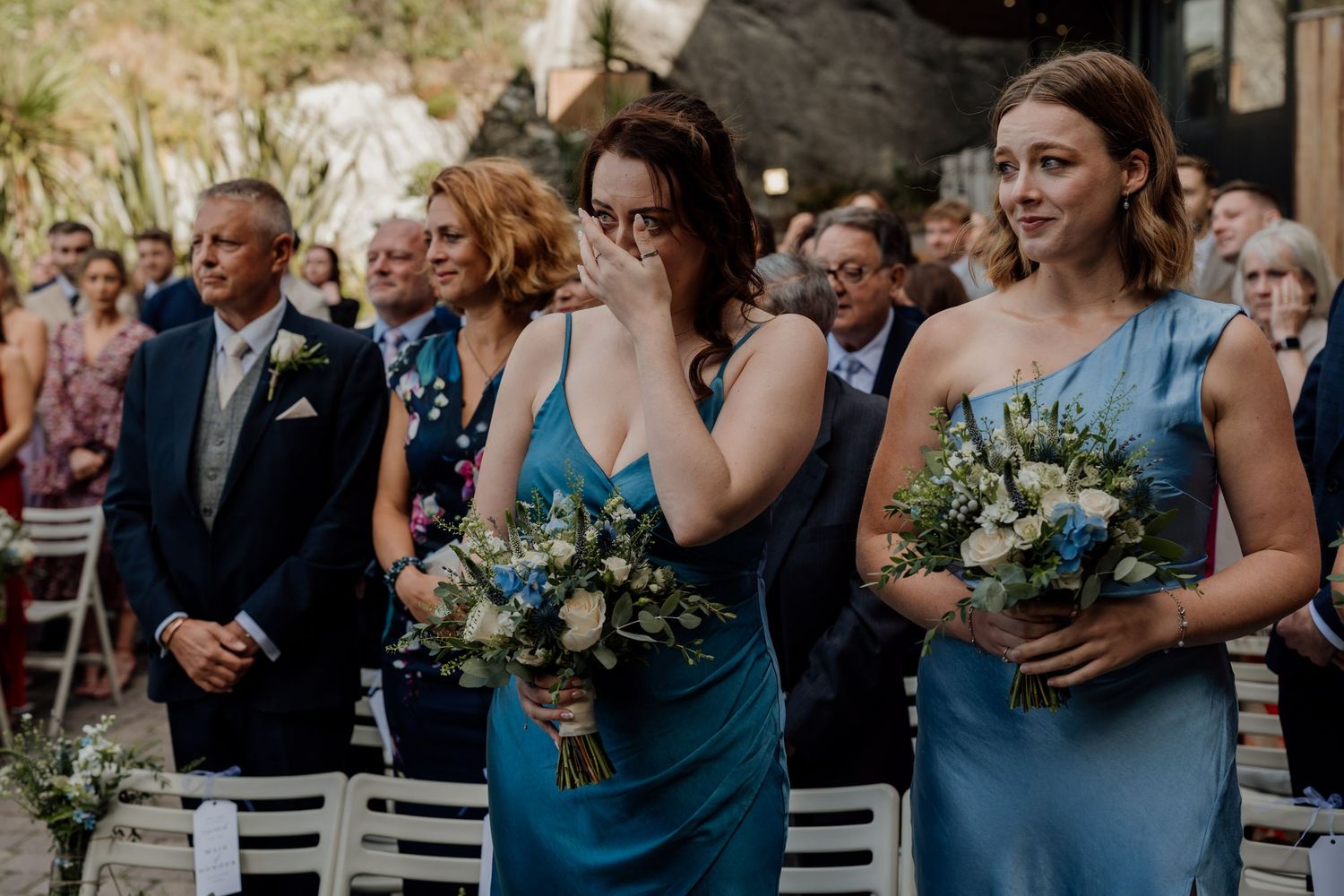 Wedding ceremony attendees seated outdoors in blue attire with floral bouquets.