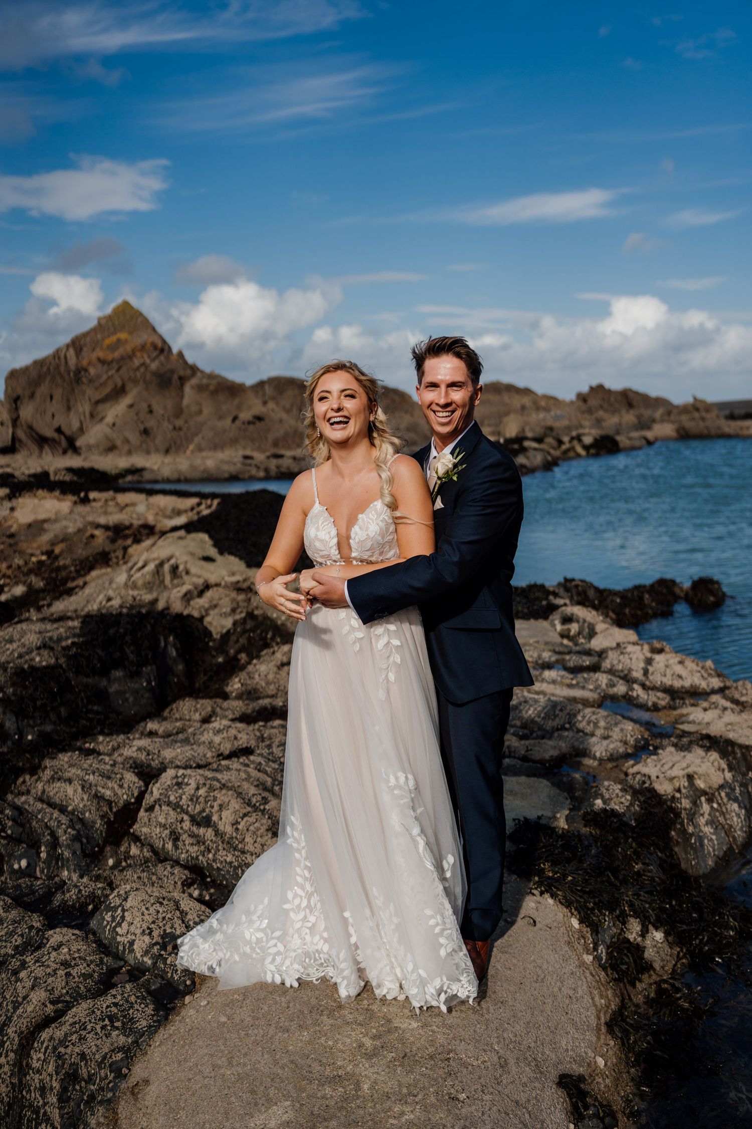Couple poses on rocky coastal formations with bright blue ocean and sky in the background.