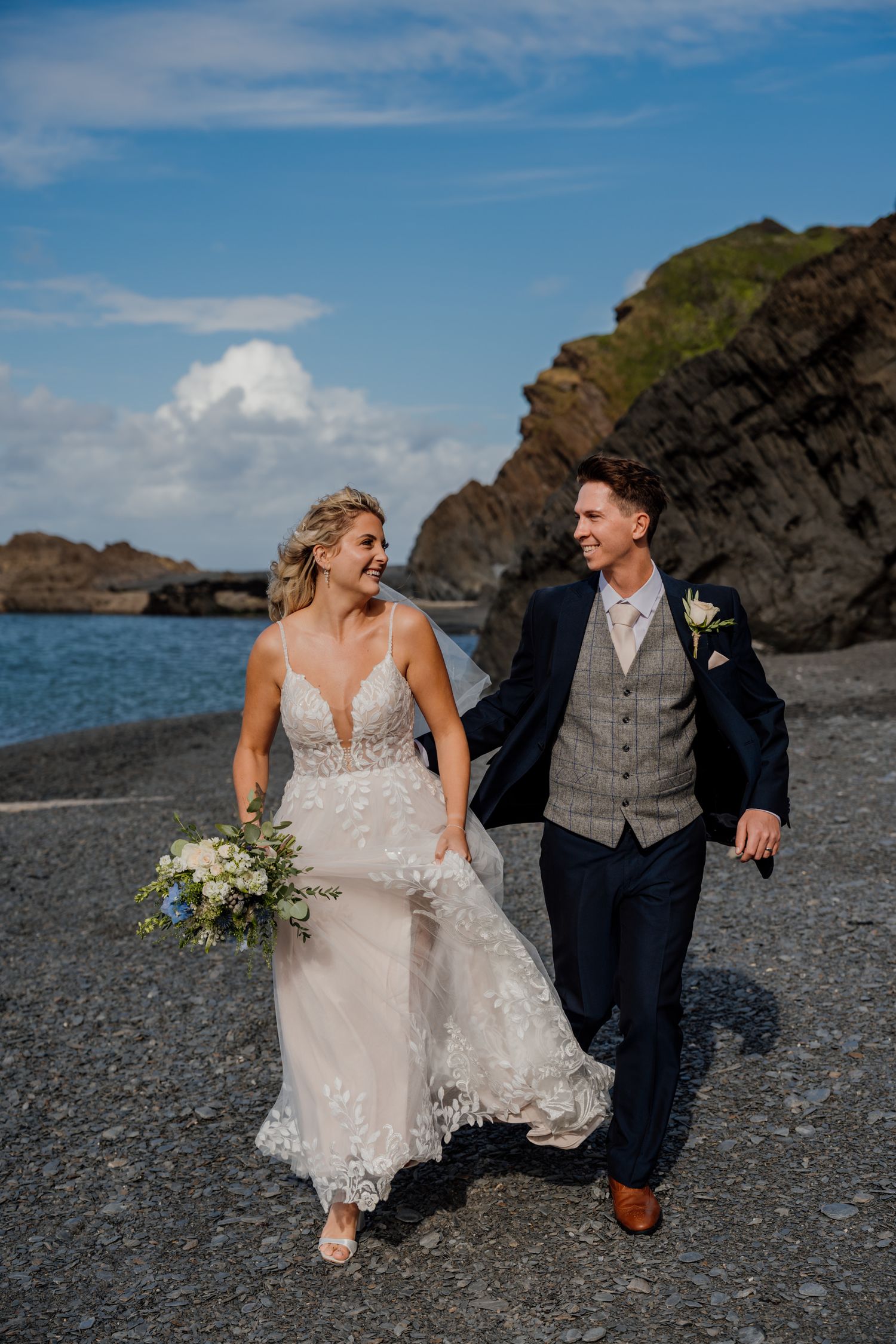 Wedding couple walking and laughing together on a pebbly beach with ocean views.