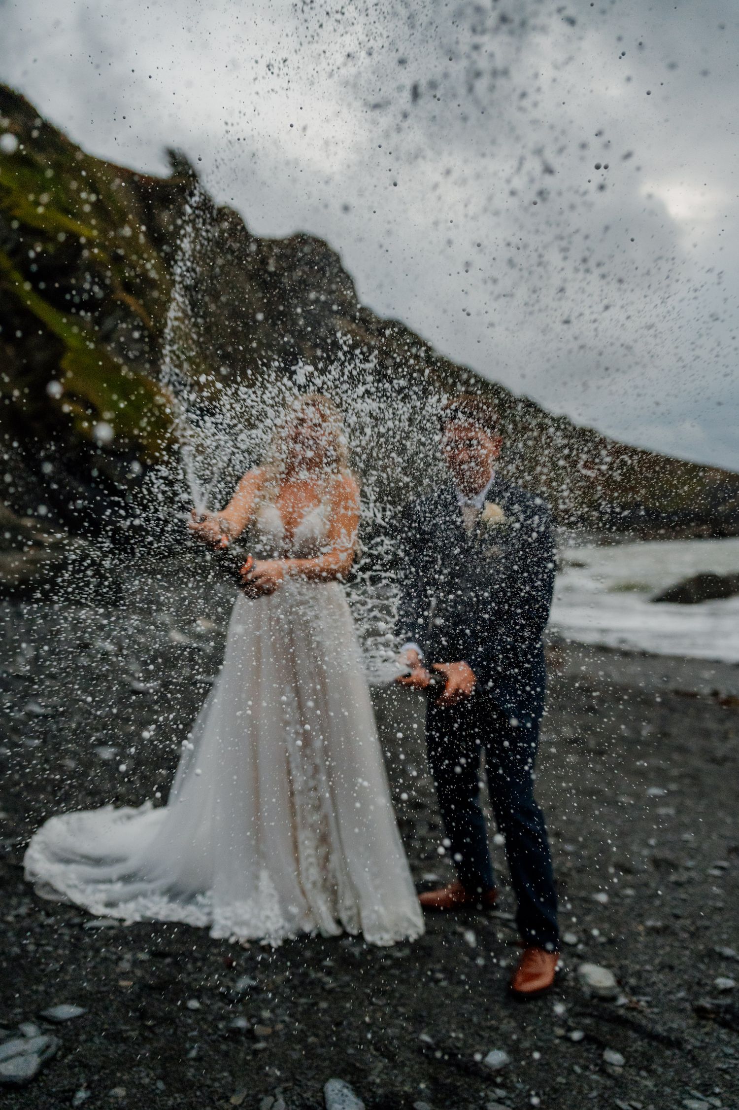 Champagne spray celebration on a moody beach with dramatic cliffs in the background.