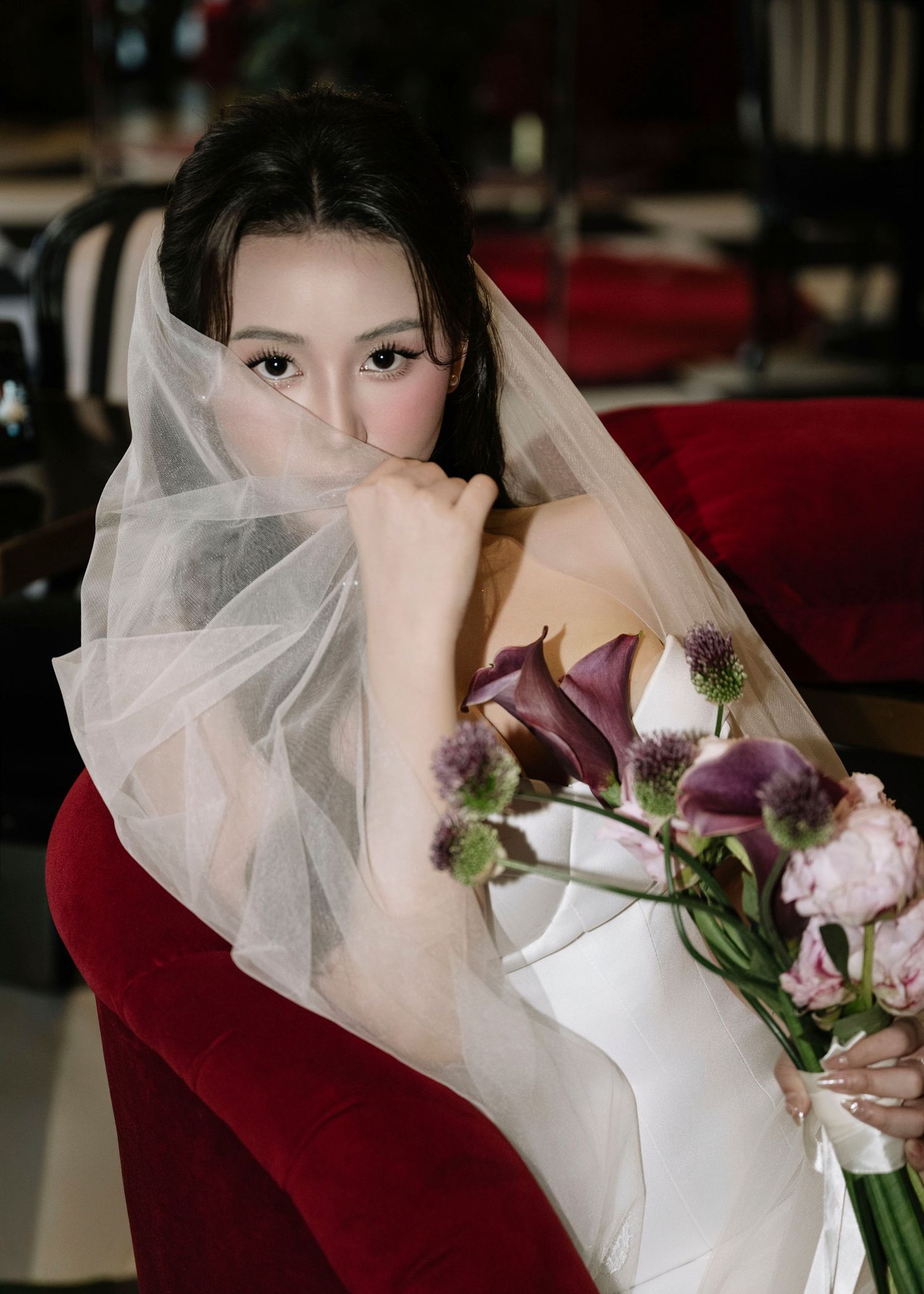 Asian bride in white strapless gown holding purple calla lily bouquet with veil draped over face.