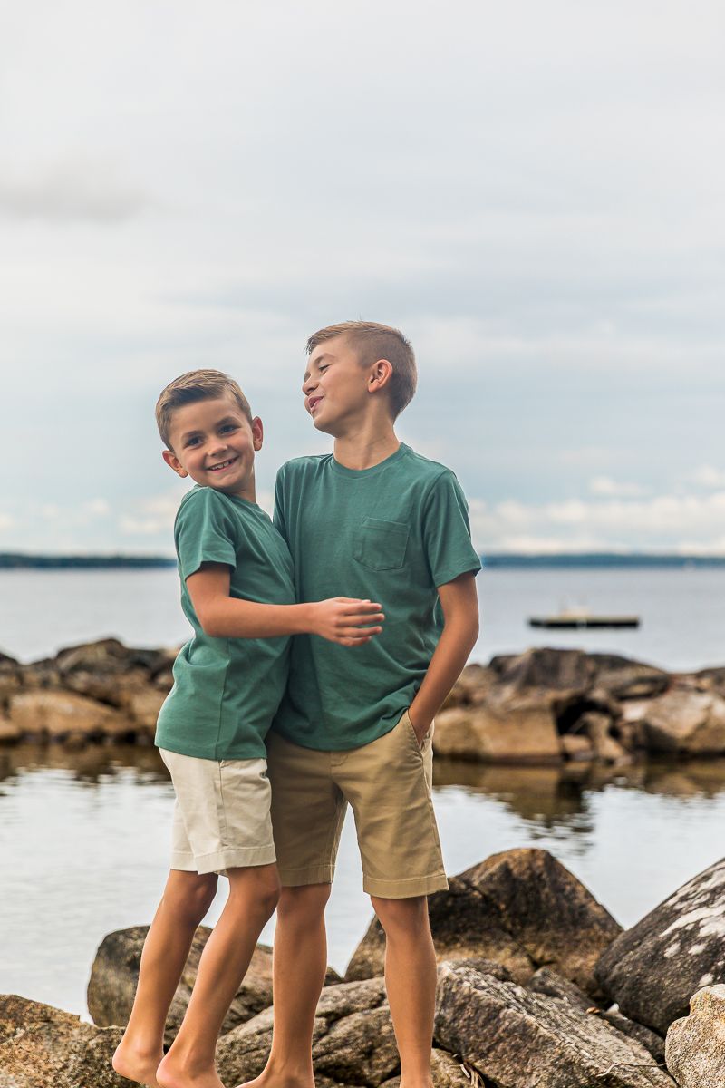 Two kids in identical outfits share a playful moment while standing on coastal rocks by the water.