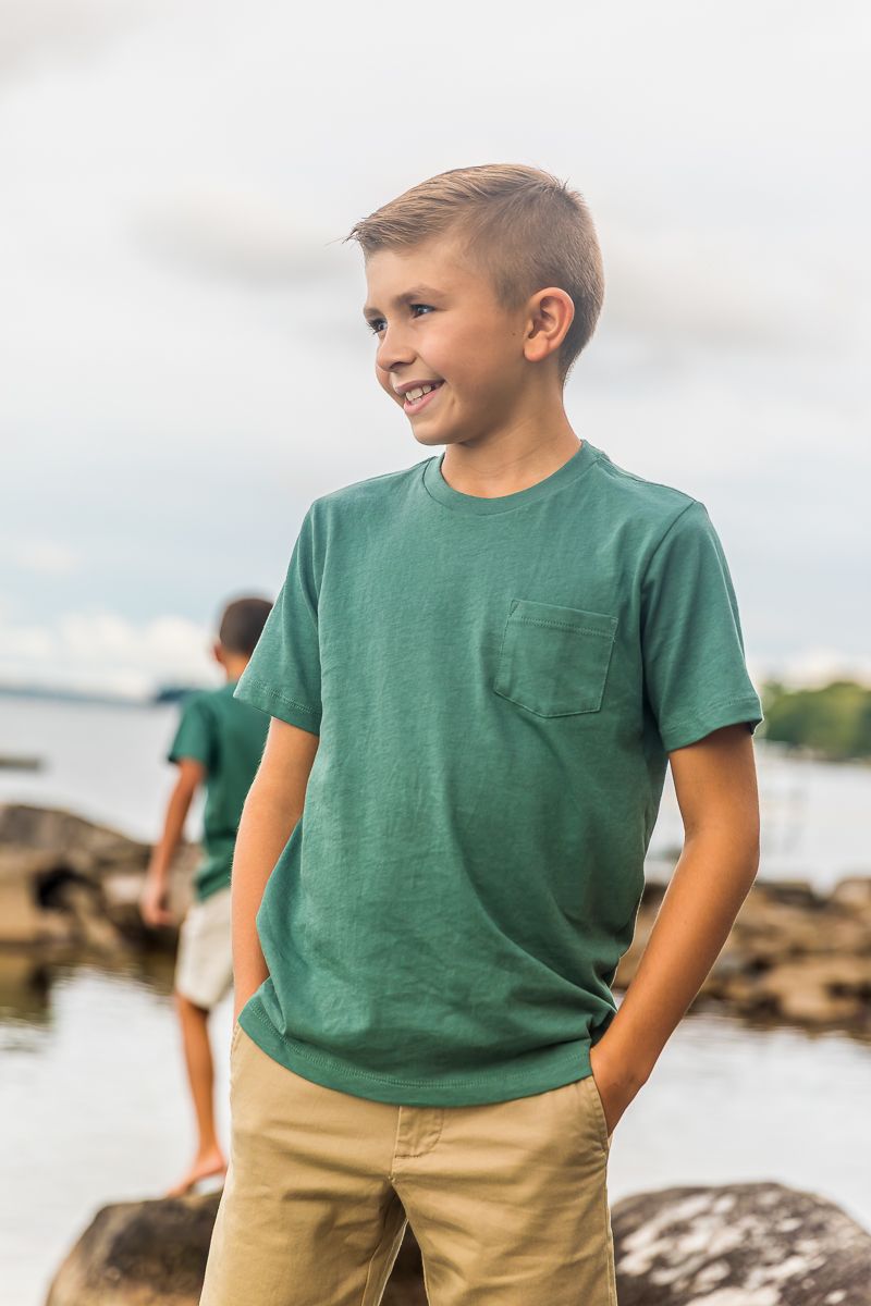 Children in matching green shirts and khaki shorts explore a rocky beach shoreline on a cloudy day.
