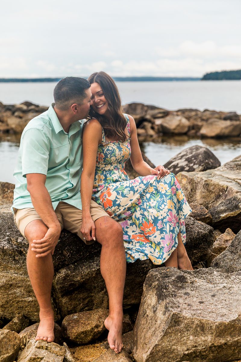 A couple sits together on rocky shoreline overlooking a serene lake, she in a floral dress and he in mint green polo.