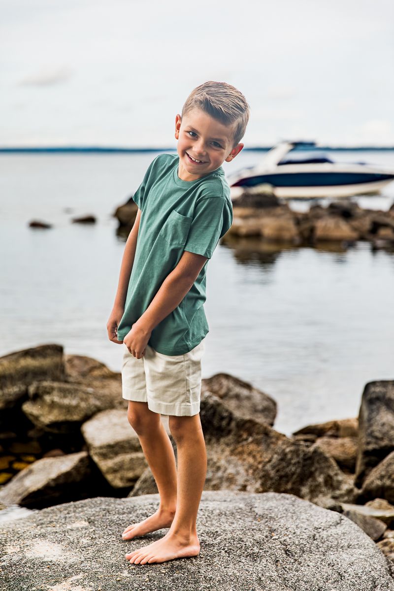 Child in a green shirt and white shorts stands barefoot on coastal rocks near calm ocean waters.