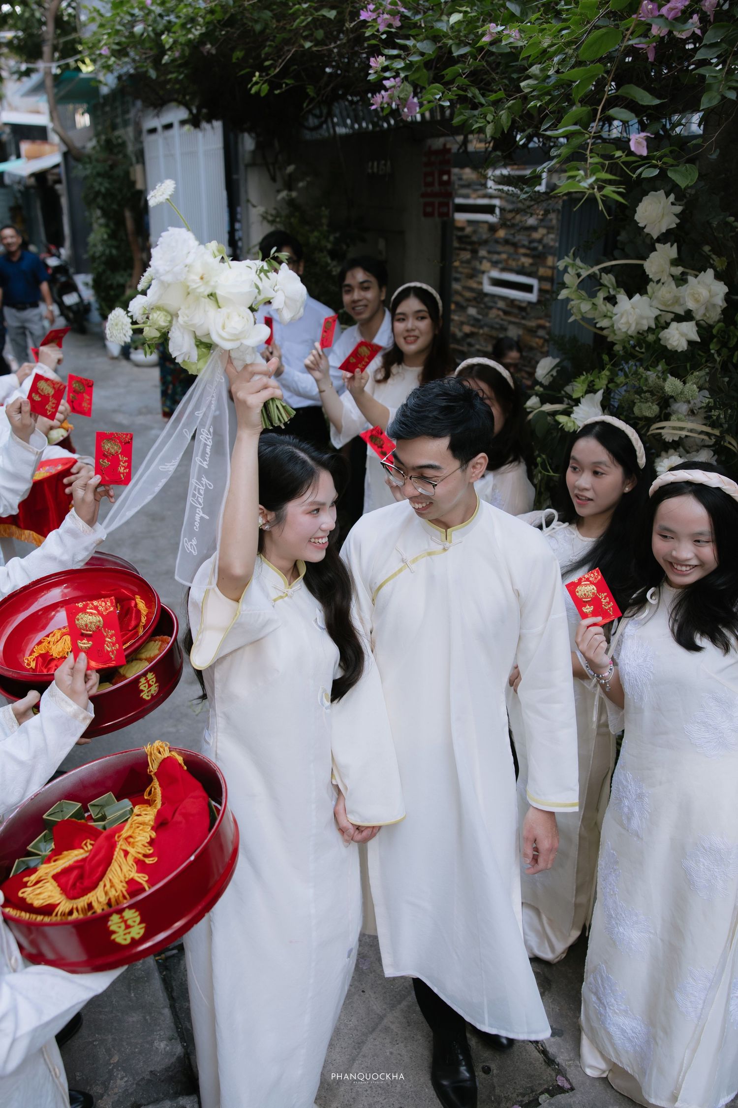 Vietnamese bride raises white bouquet while walking with groom through traditional wedding procession outdoors.