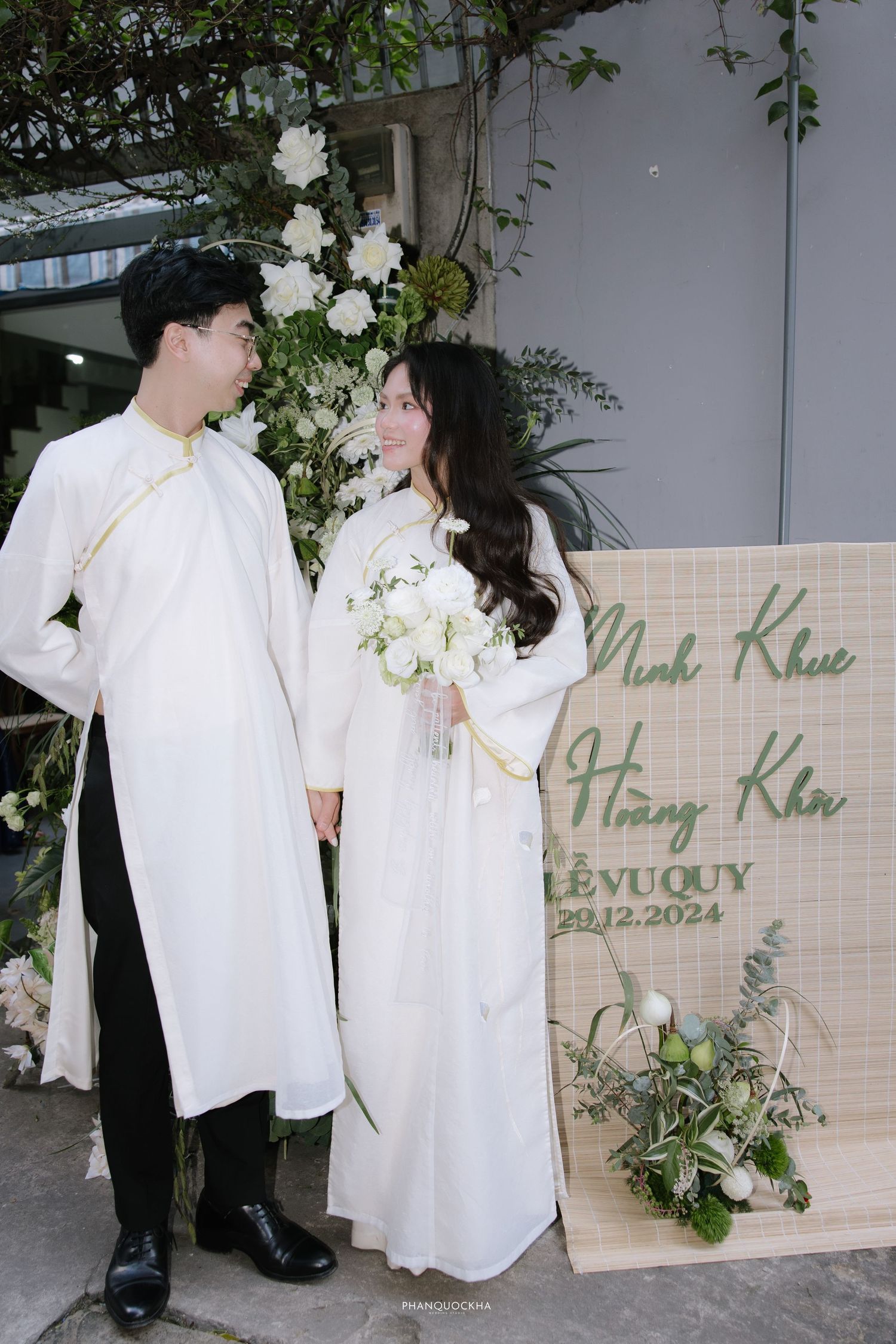 Vietnamese bride holds white bouquet while groom gazes at her lovingly at their December 2024 wedding.