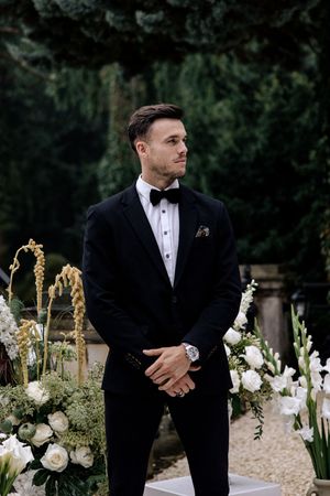 A groom in a classic black tuxedo poses by elegant white floral arrangements in an outdoor garden setting.