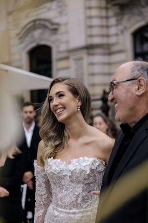 A woman in a white off-shoulder wedding dress smiles joyfully while conversing with someone in dark formal attire.