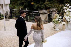 A wedding couple holds hands and walks together on a gravel path surrounded by romantic garden decor and flowers.